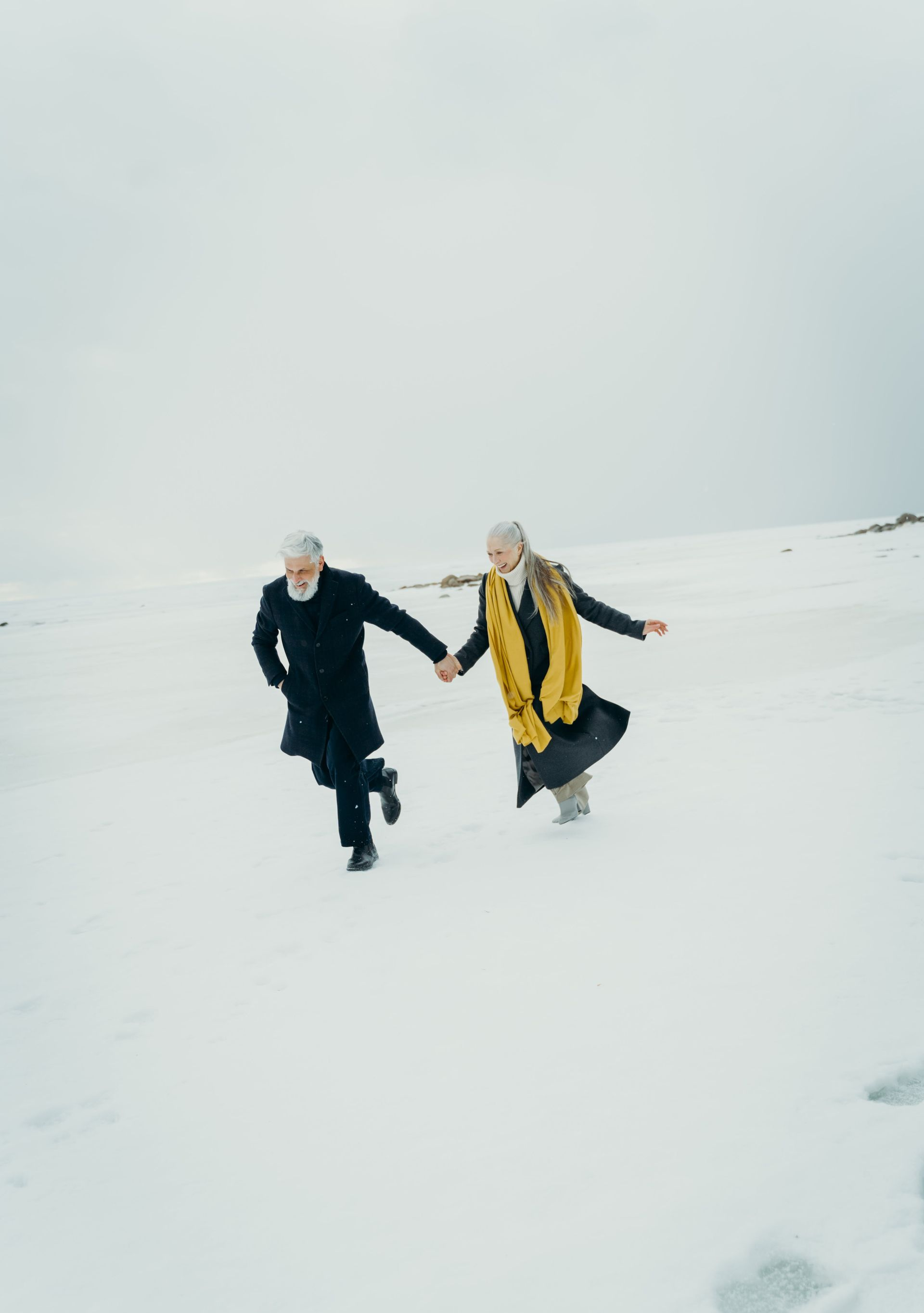 Two people run hand-in-hand in a snowy field. One wears a black coat, the other, a yellow scarf. Overcast sky.