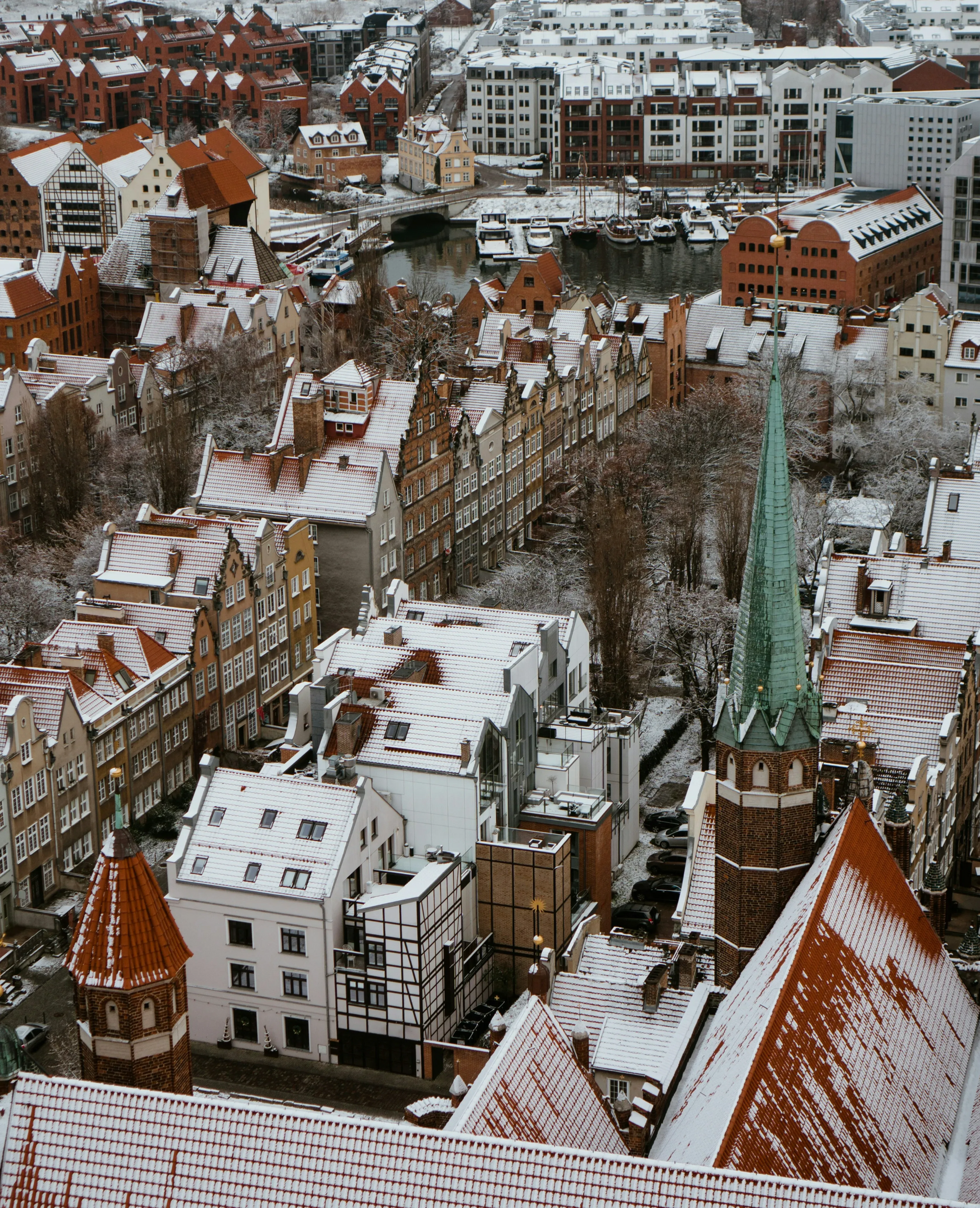 Overhead view of a snow-covered European city with red rooftops and a green-spired church.