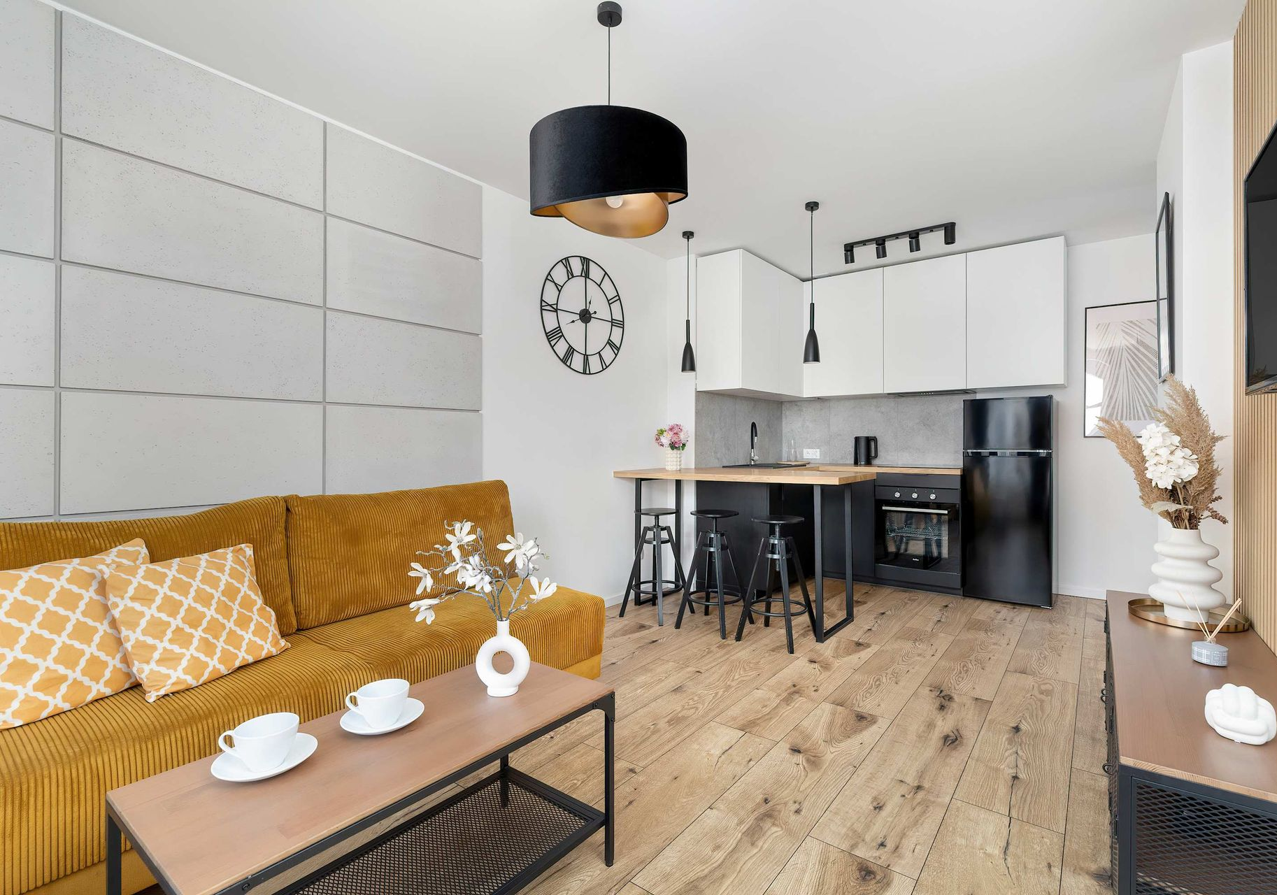 Nowoczesne studio z musztardowo-żółtą sofą, drewnianym stolikiem kawowym, blatem kuchennym i czarnymi sprzętami kuchennymi.
