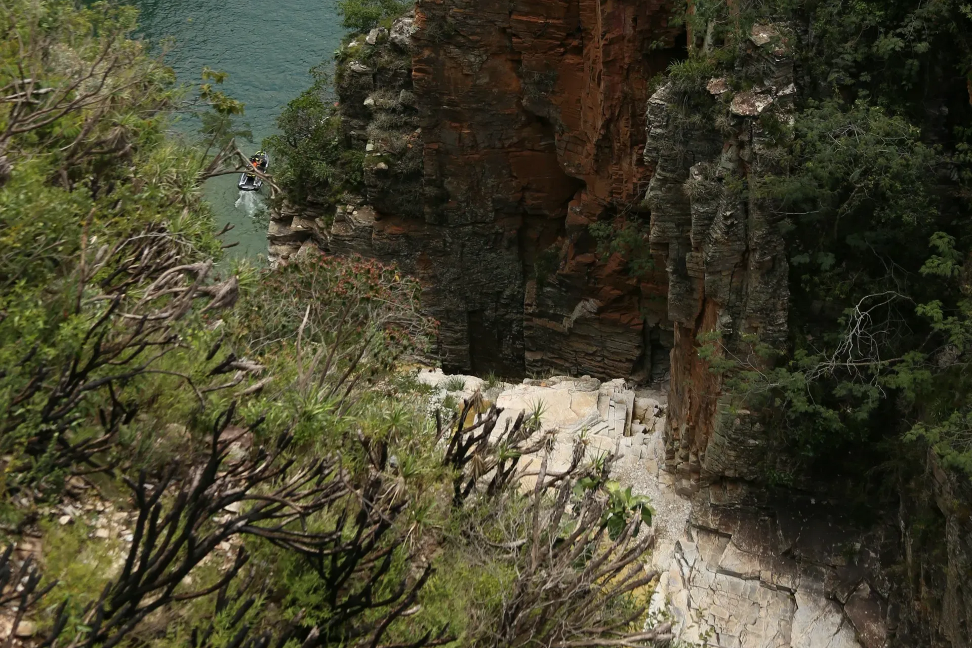 Canyon with green water, boats, and people on a cliffside, surrounded by trees.