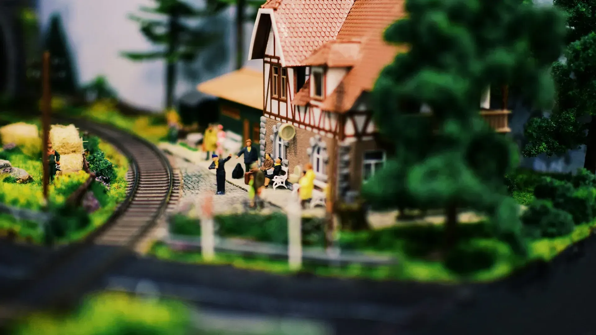Miniature train station with tiny figures, tracks, and a red-roofed building, set outdoors.