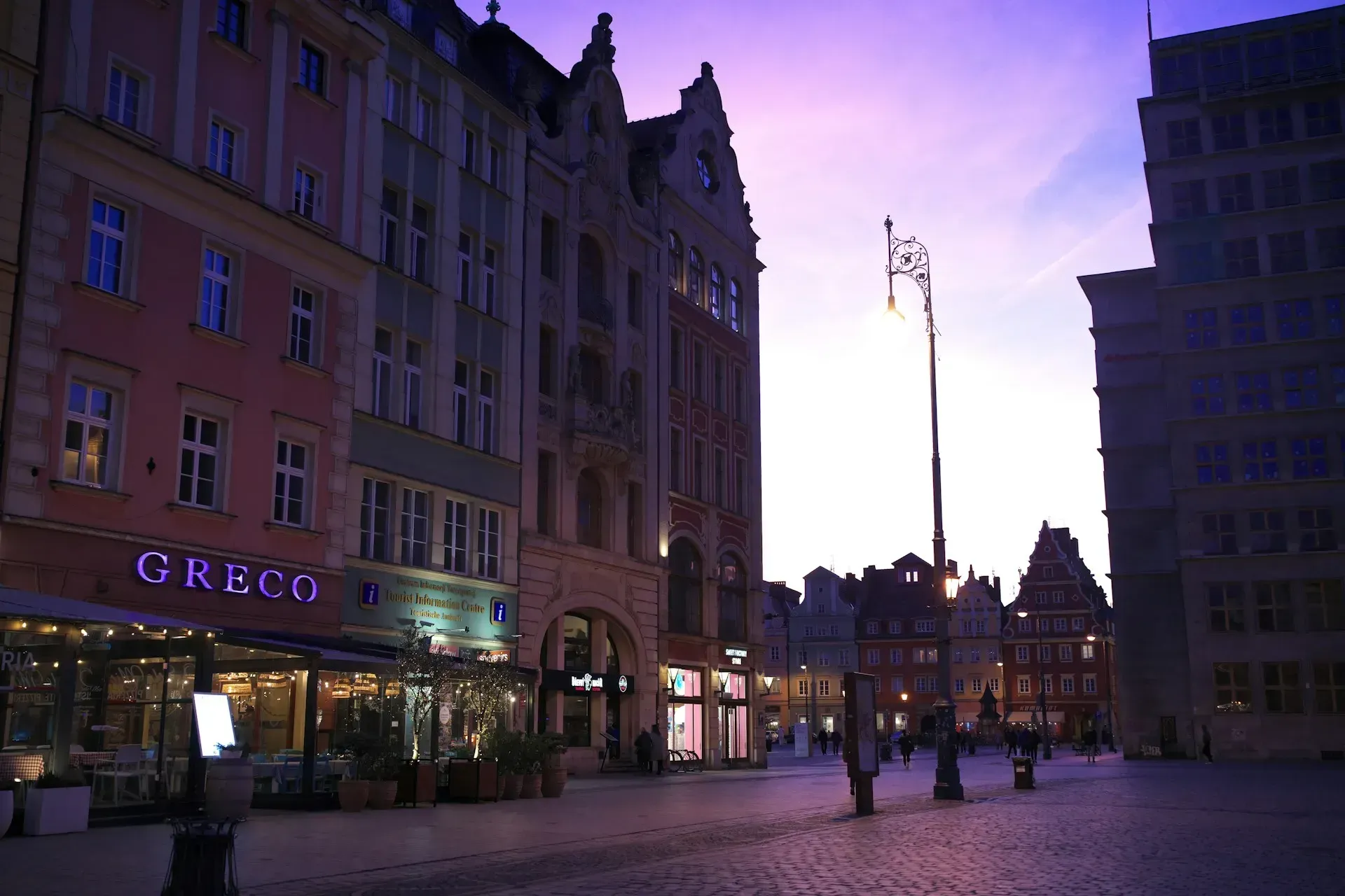 Europejski plac miejski o zmierzchu, kolorowe budynki, latarnie uliczne, delikatne fioletowe niebo.