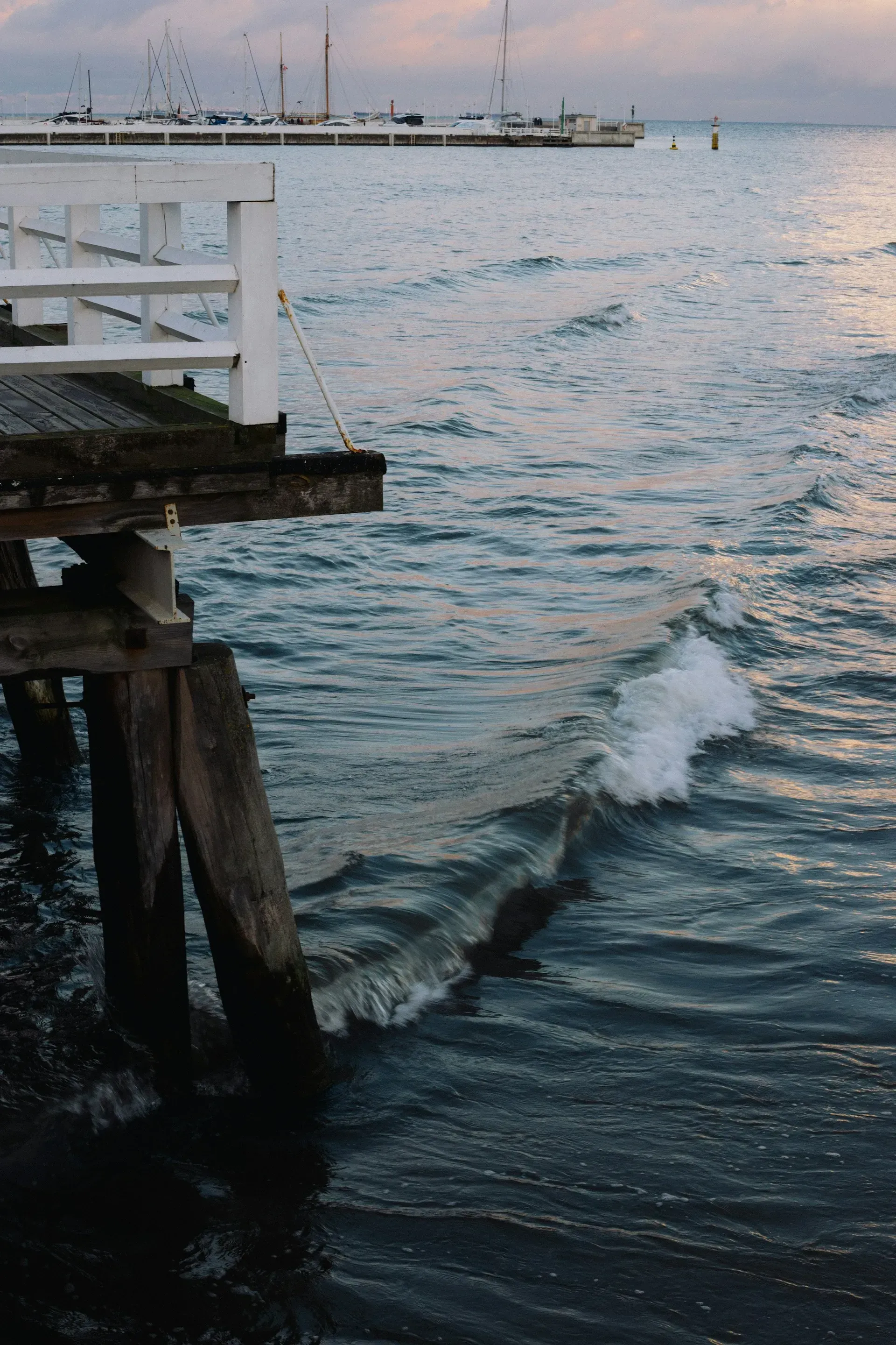 Fale rozbijają się o drewniane pale mola; spokojny ocean, w oddali widać łodzie, a niebo jest zachmurzone.