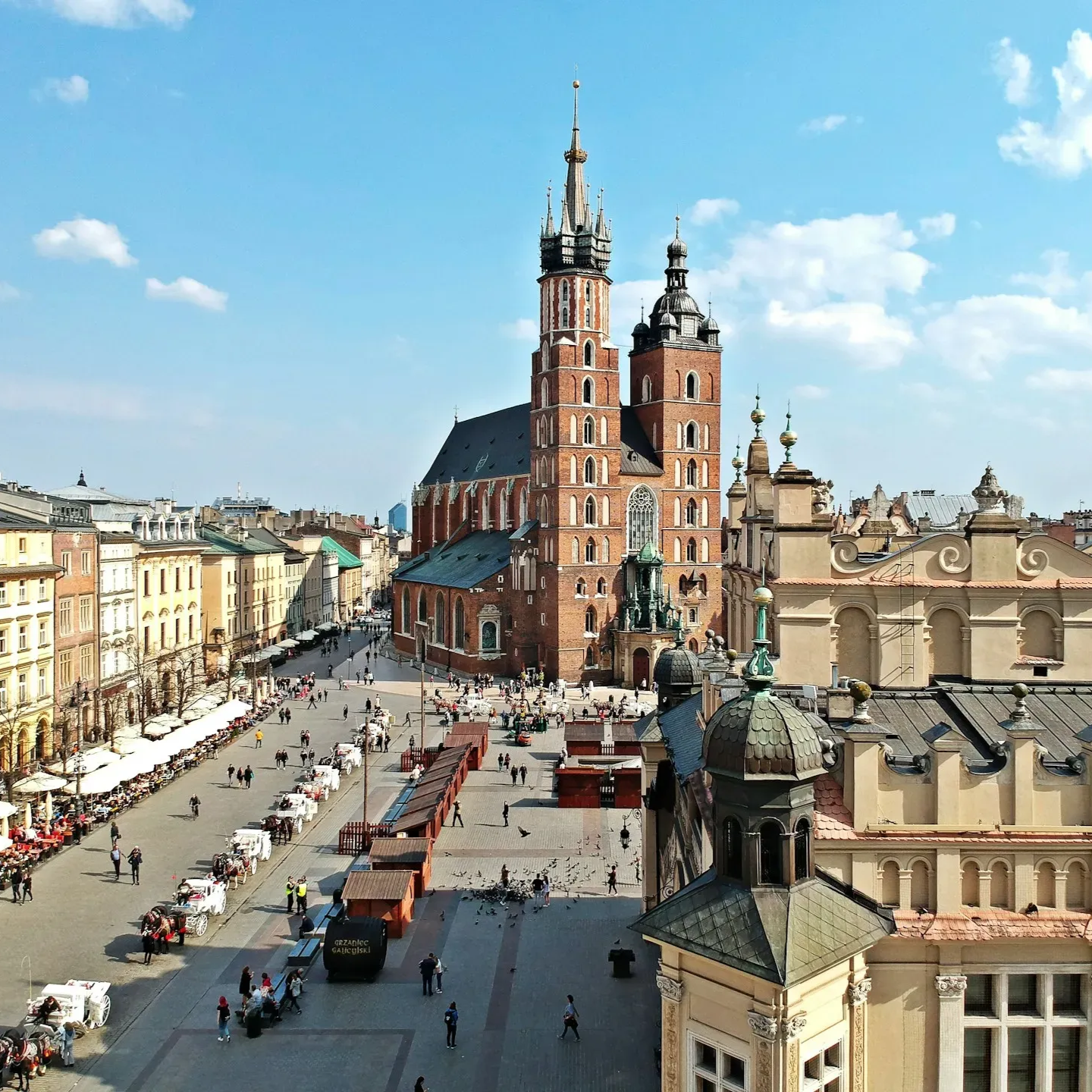 Rynek Główny w Krakowie z Bazyliką Mariacką i Sukiennicami w słoneczny dzień; spacerujący ludzie.