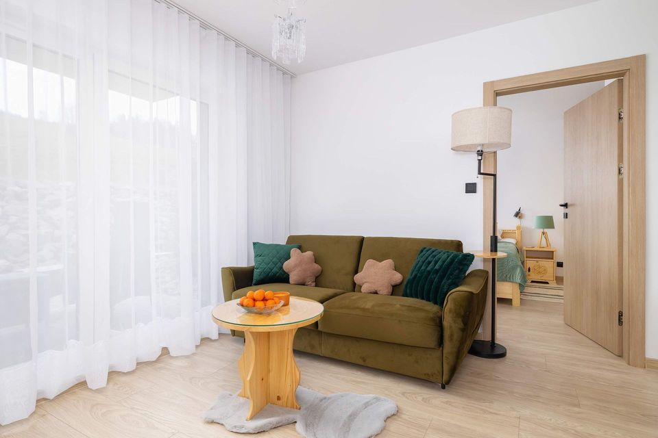 Jasny salon z oliwkową sofą, lampą podłogową, okrągłym stołem i cienkimi zasłonami przy dużym oknie
