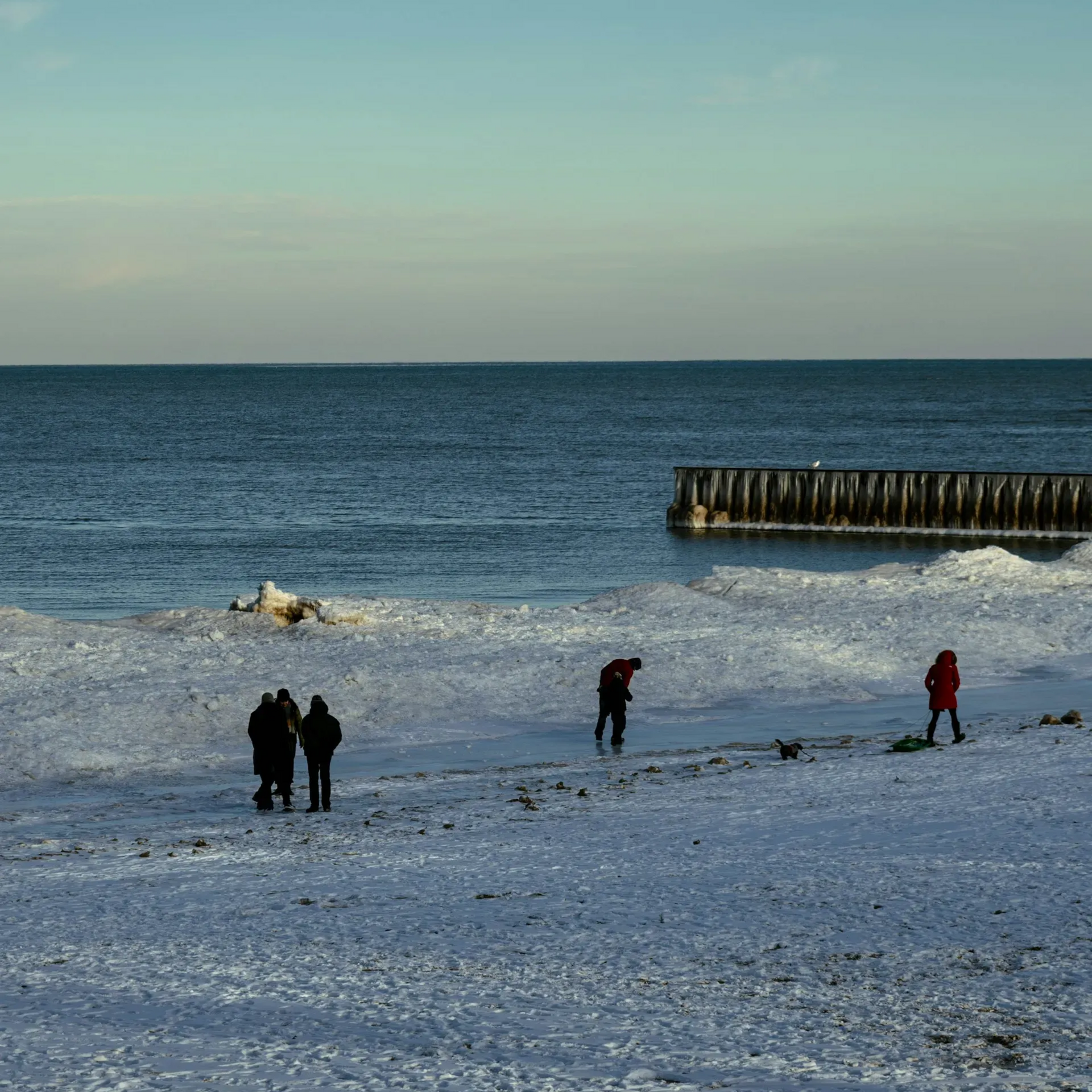 Ludzie na zaśnieżonej plaży w pobliżu lodowatej wody i drewnianego falochronu; pochmurny dzień.