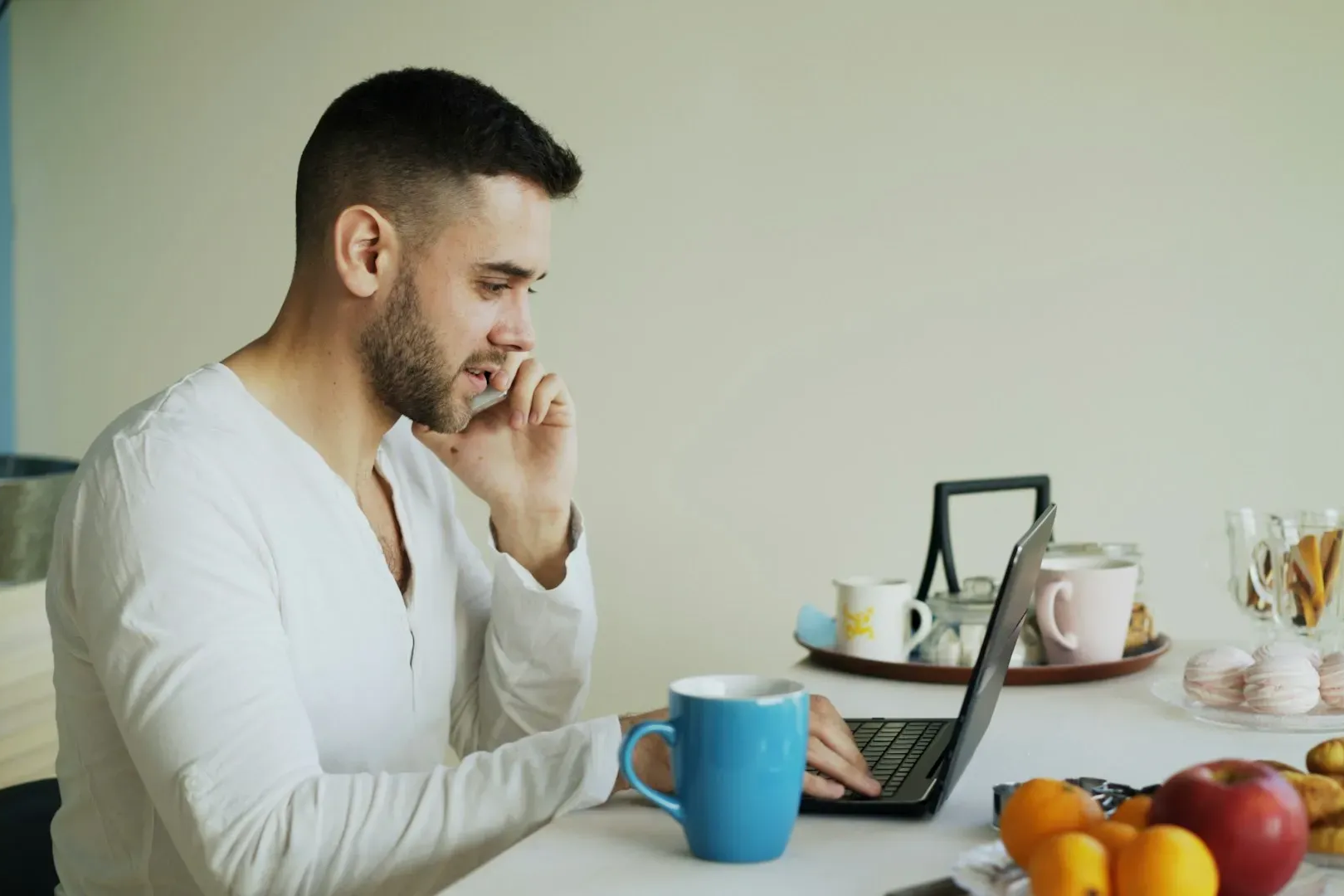 Man in white shirt at table with laptop, blue mug, fruit; talking on phone.