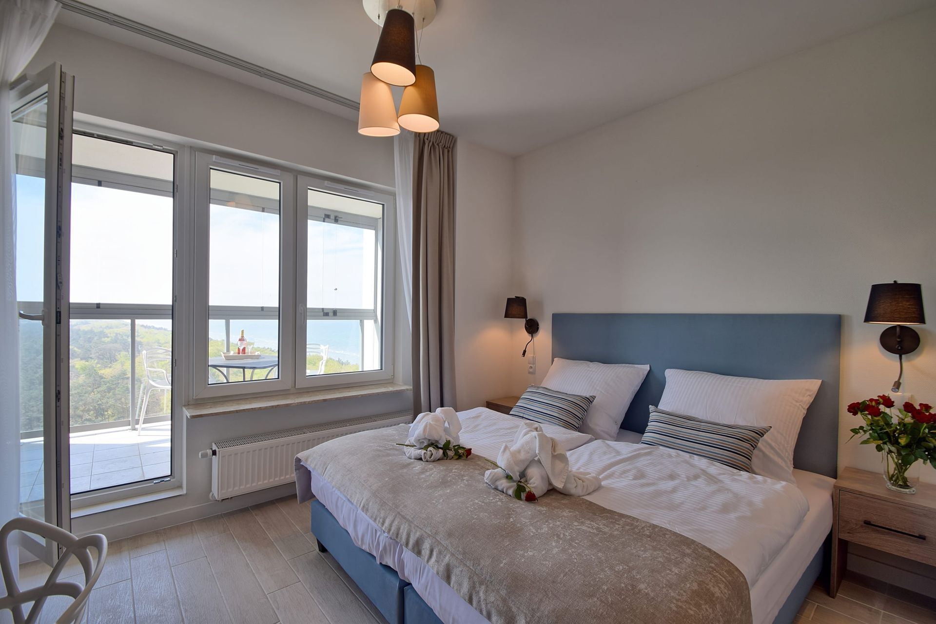 Przytulna, jasna sypialnia z dużym łóżkiem typu queen-size, miękkim, niebieskim zagłówkiem, dużymi oknami i widokiem z balkonu.