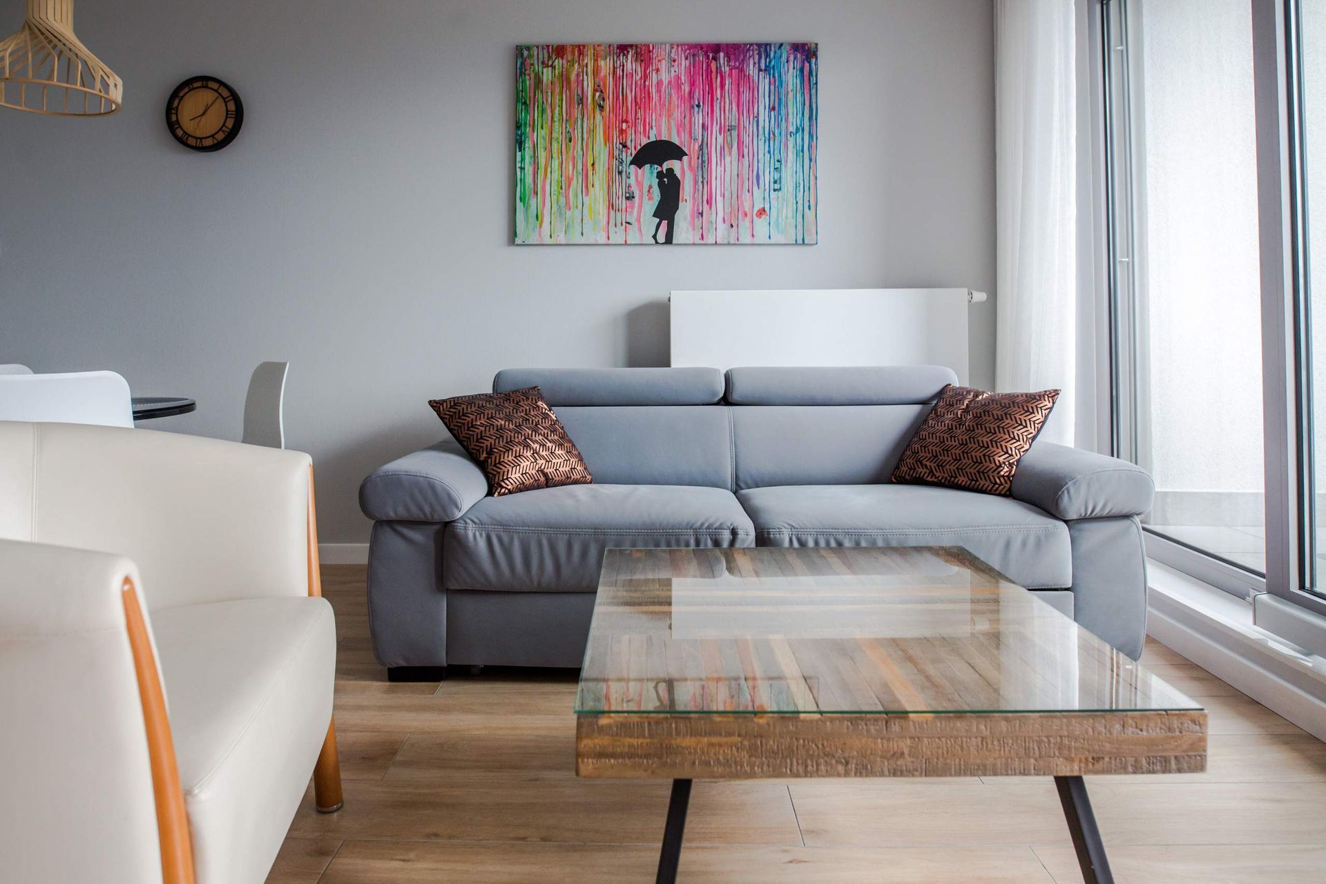 Nowoczesny salon z szarą sofą, drewnianym stolikiem kawowym ze szklanym blatem i kolorową, abstrakcyjną sztuką na szarej ścianie.