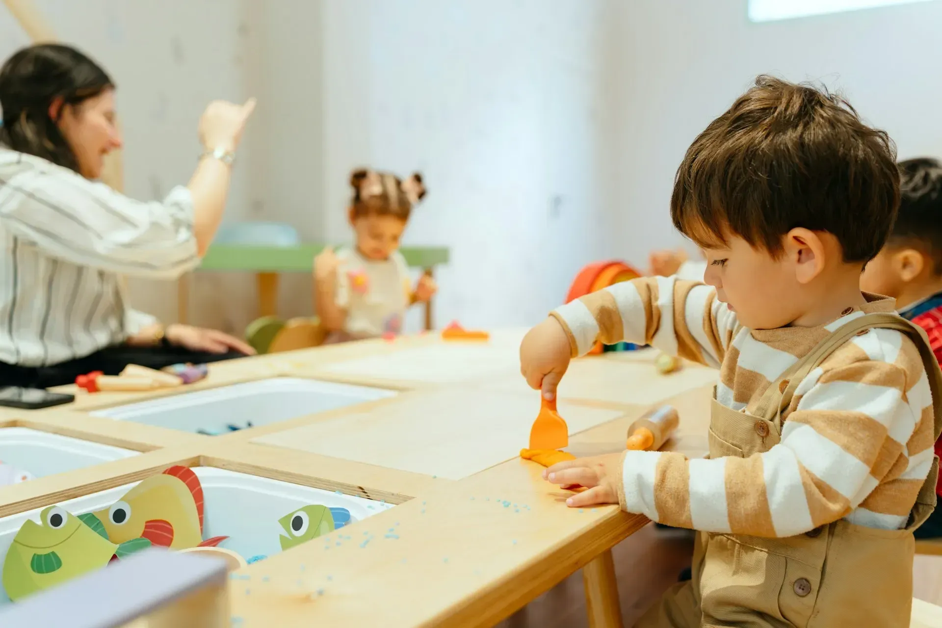 Boy in striped shirt playing with toys at a table in a playroom; teacher and other children nearby.