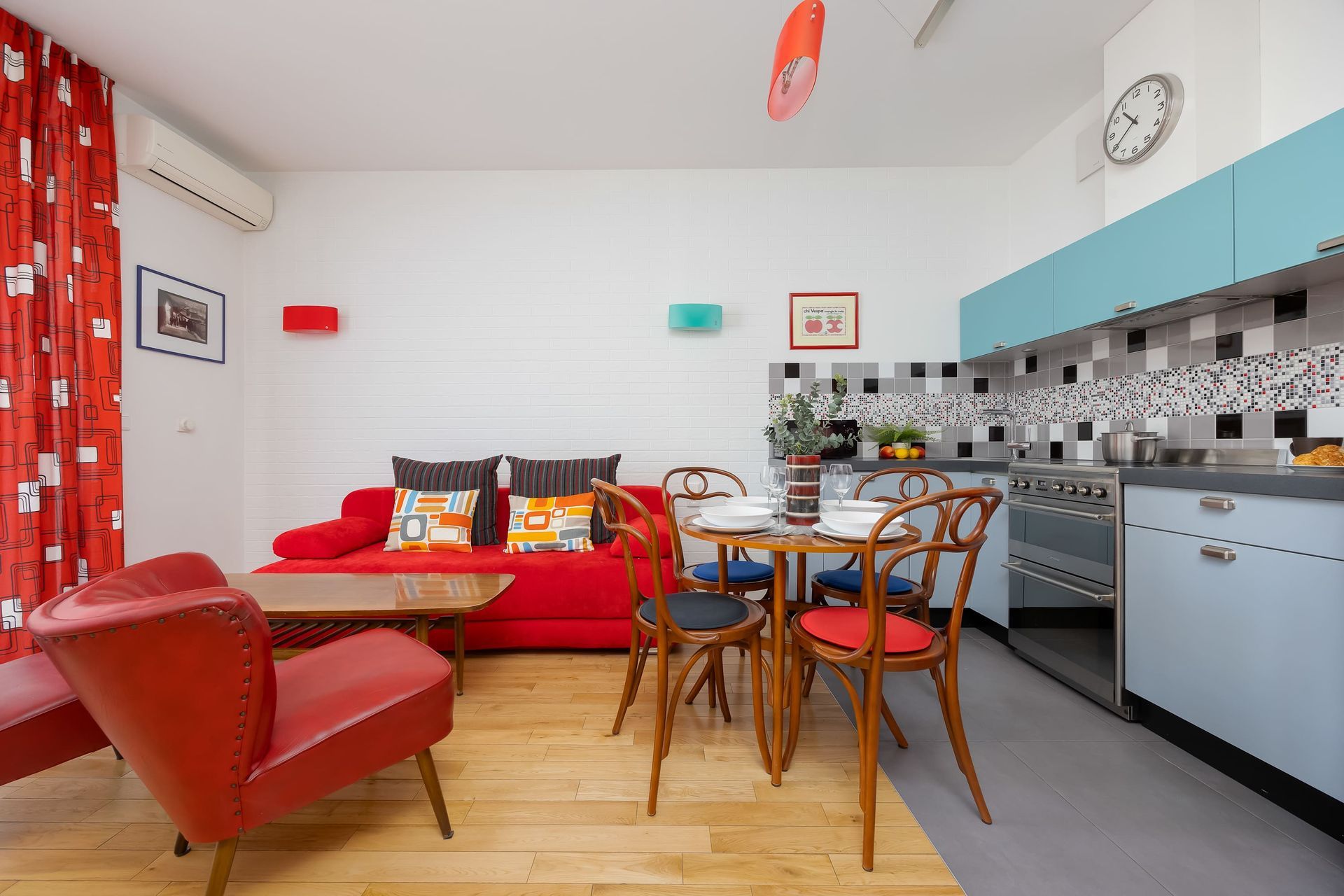 Jasne mieszkanie z czerwoną sofą i fotelem, niewielkim zestawem jadalnym oraz niebieską kuchnią ze wzorzystymi płytkami na ścianie nad blatem.