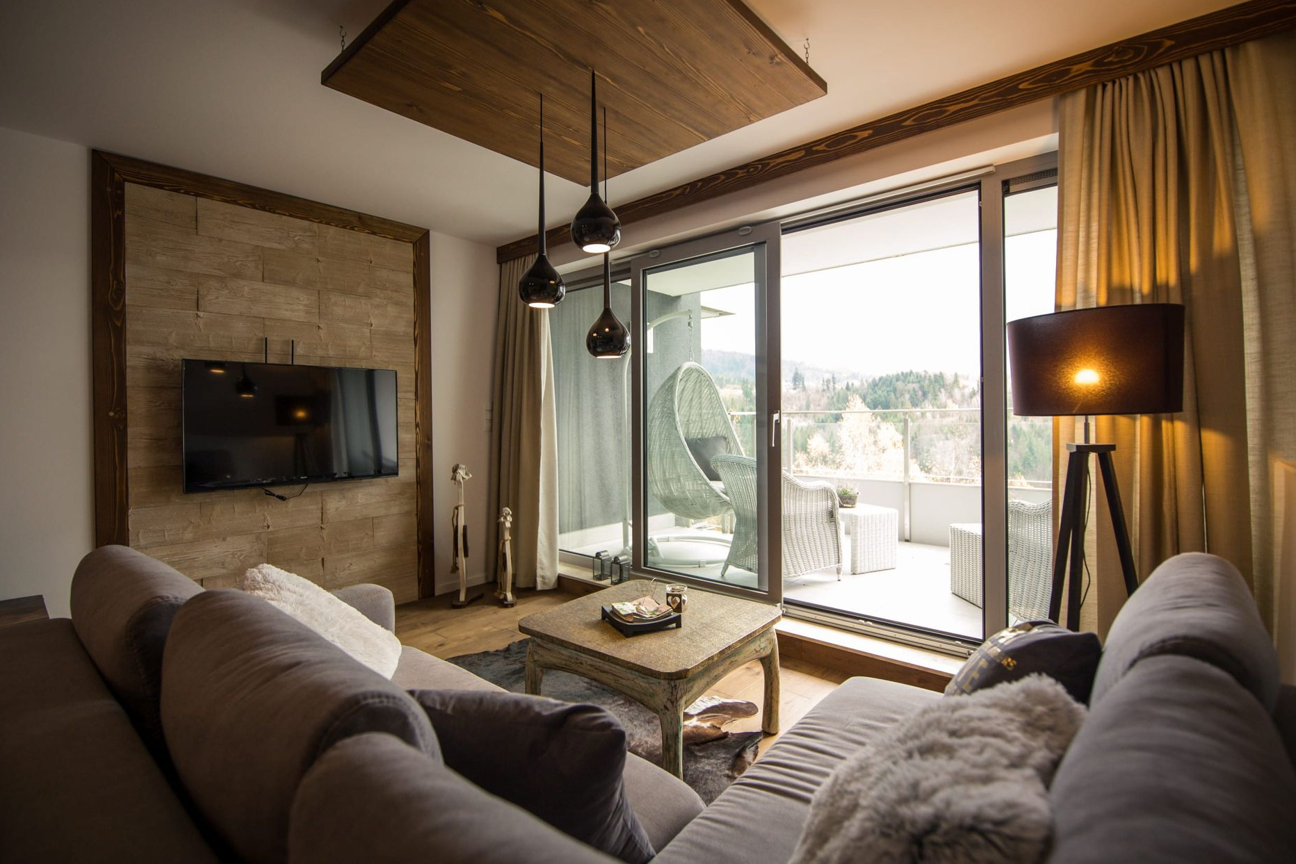 Salon z dużą, szarą sofą, telewizorem na ścianie o ciekawej fakturze i przesuwanymi drzwiami na balkon.