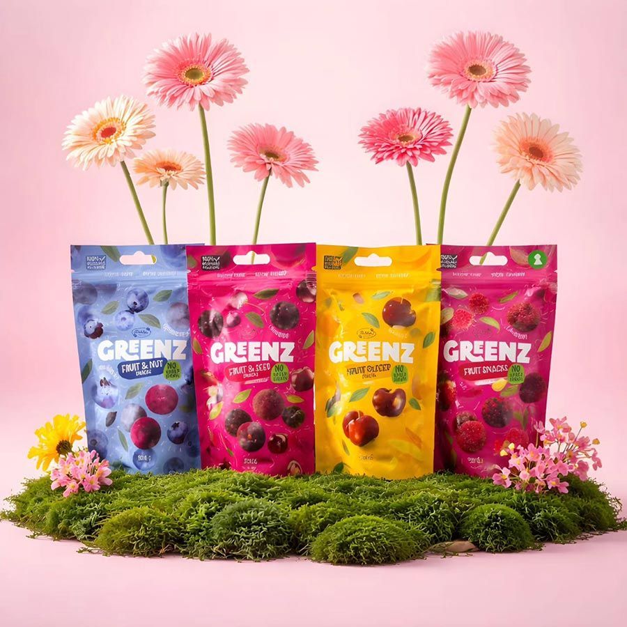 Cuatro bolsas de snacks de fruta GRENZ de colores, sobre musgo con flores rosas y un fondo rosa