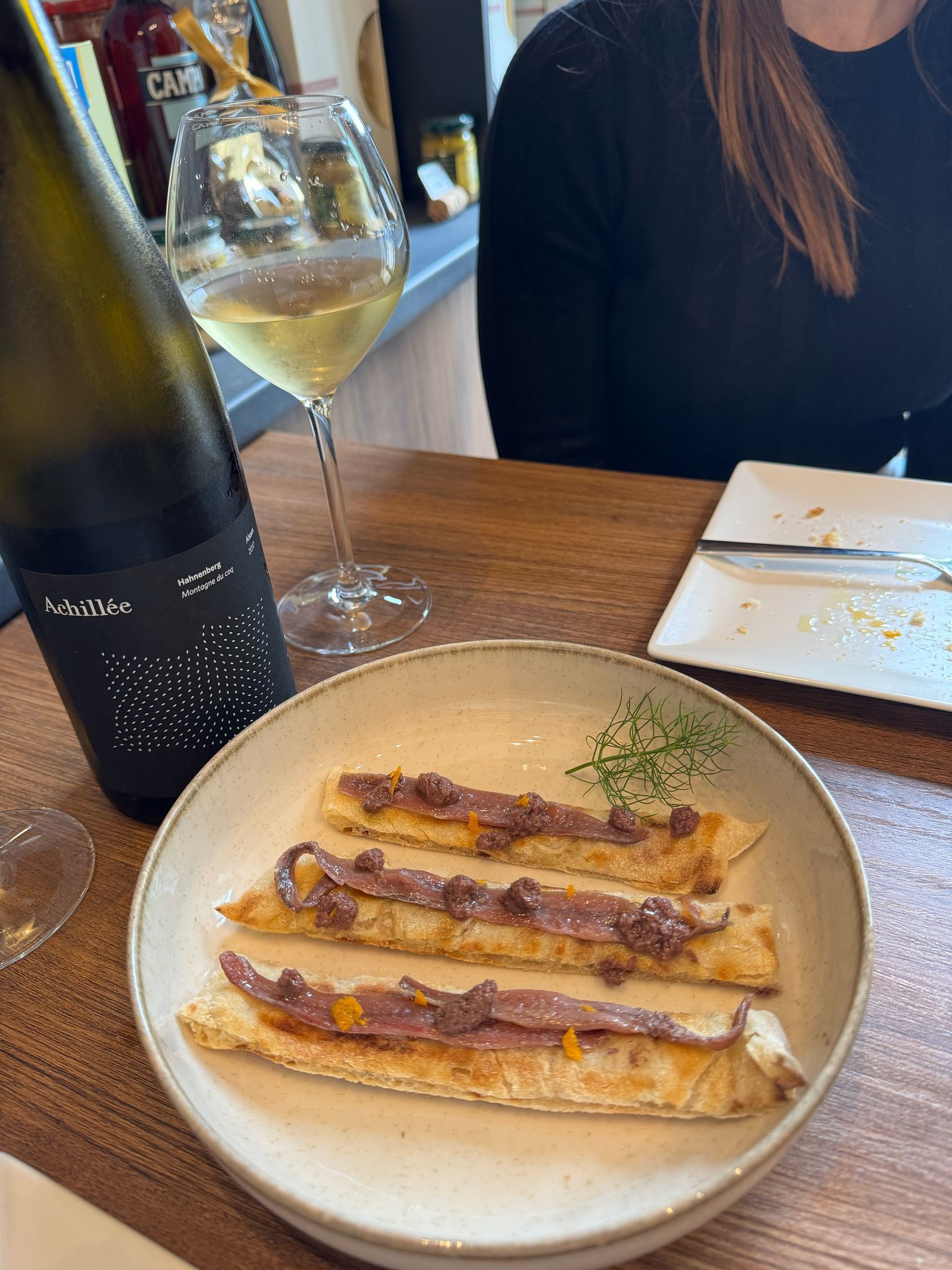Una mujer está sentada en una mesa con un plato de comida y una copa de vino.