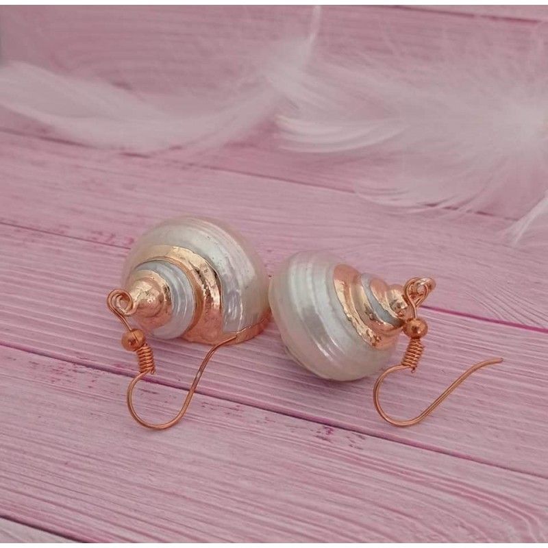Des boucles d'oreilles en forme de coquillage, ornées de cuivre, reposent sur une surface en bois rose. Des plumes blanches se dessinent en arrière-plan.