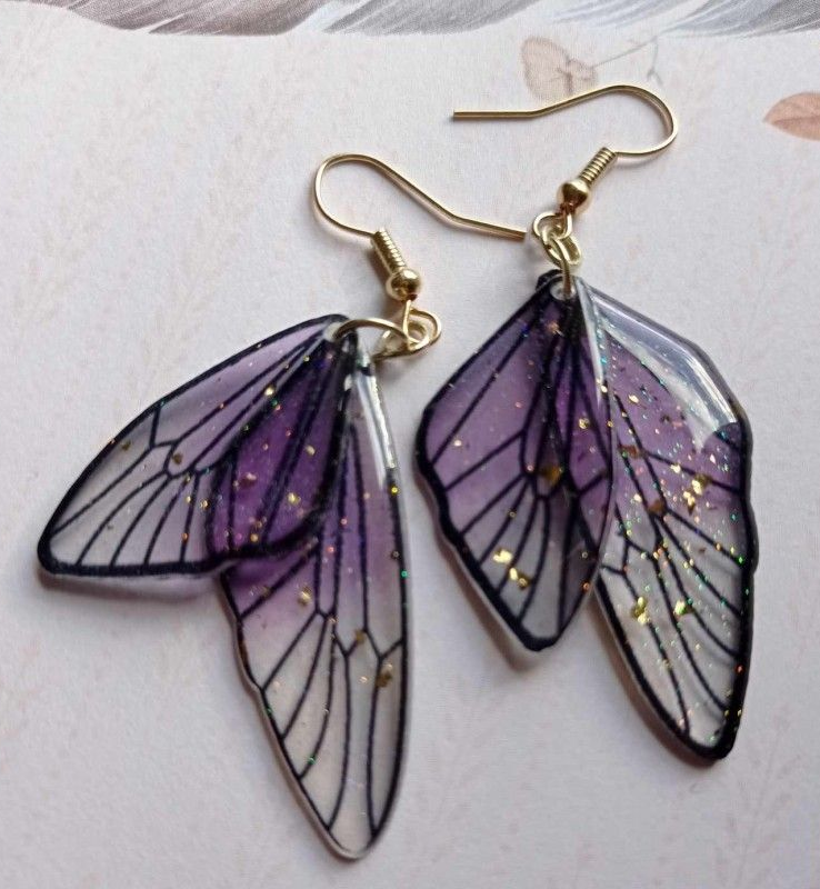 Boucles d'oreilles ailes de papillon violettes et blanches avec détails et crochets dorés.