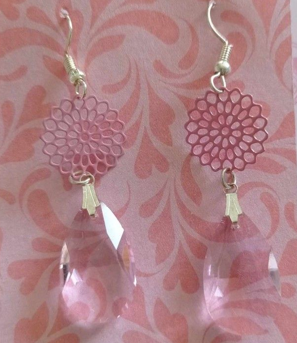 Boucles d'oreilles roses ornées de motifs floraux en filigrane et de pampilles en cristal en forme de larme, sur un fond rose à motifs.