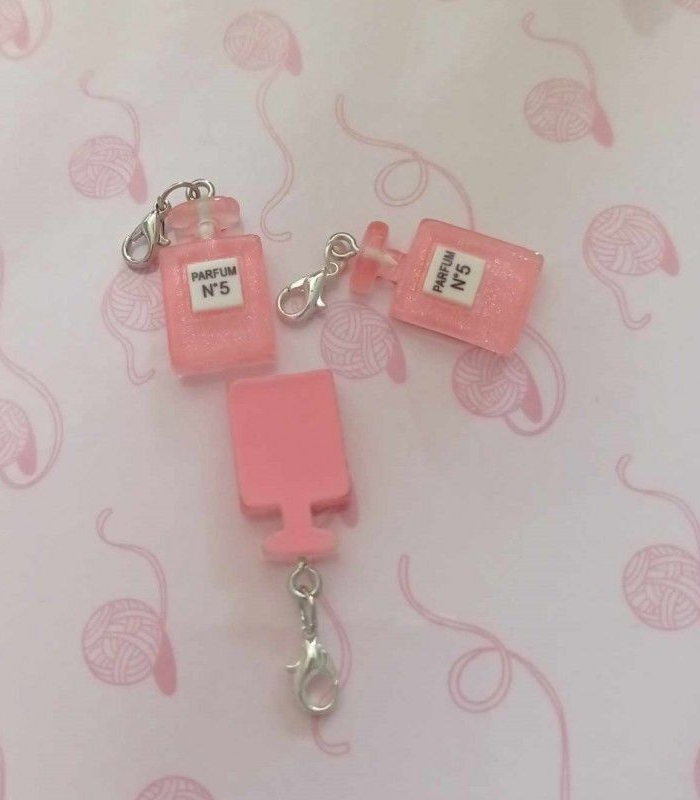 Trois breloques en forme de flacons de parfum roses avec fermoirs argentés sur fond rose.