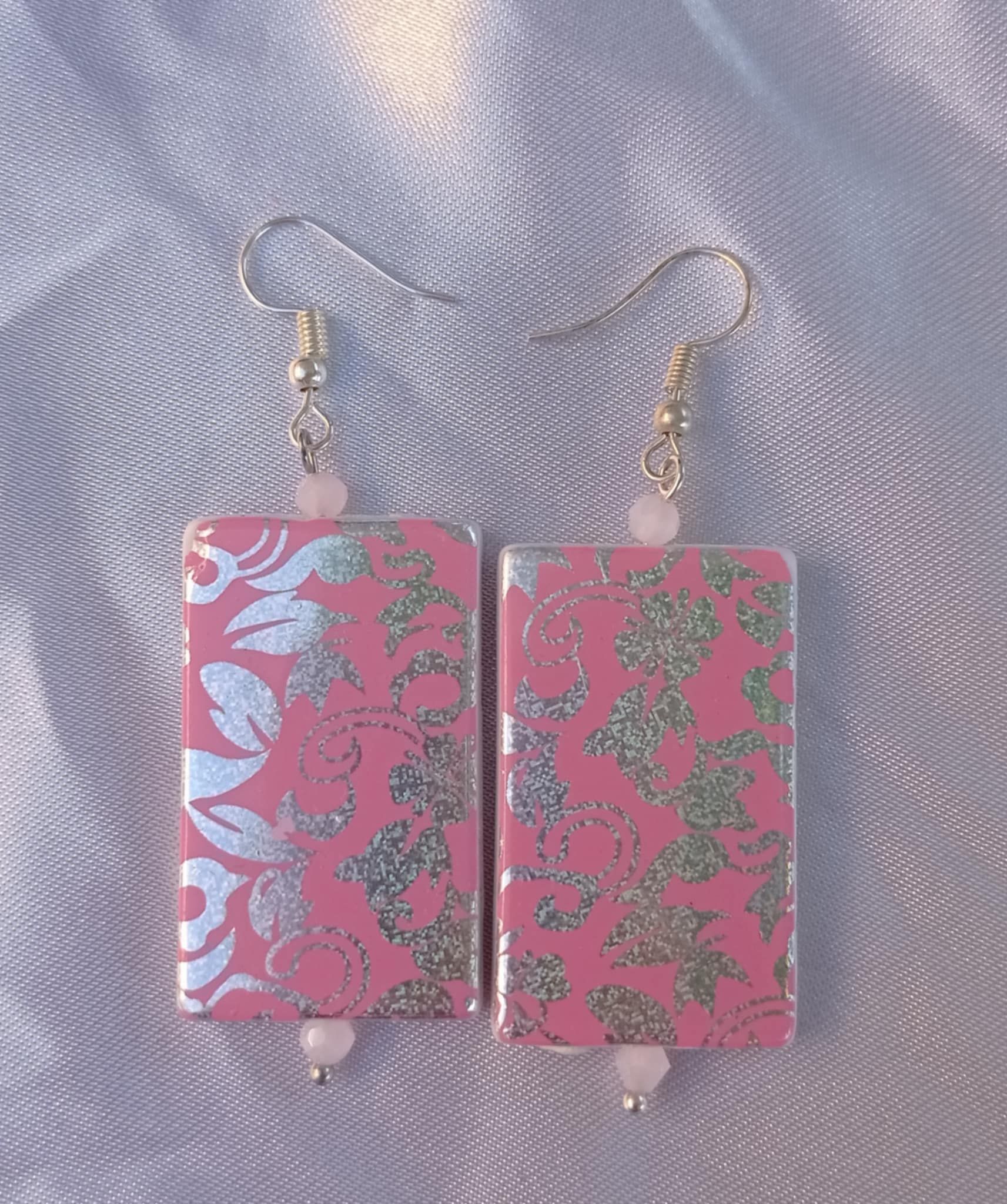 Boucles d'oreilles rectangulaires roses avec motif floral argenté, perles blanches et crochets argentés.