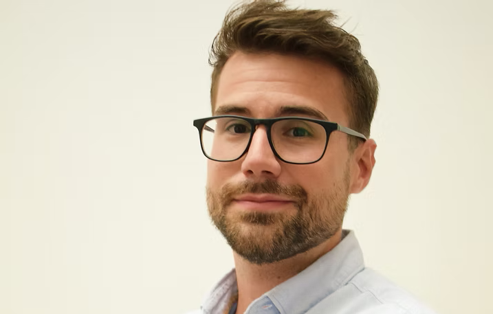 Hombre con cabello castaño corto y gafas sonriendo, vistiendo una camisa azul claro, sobre un fondo blanco.