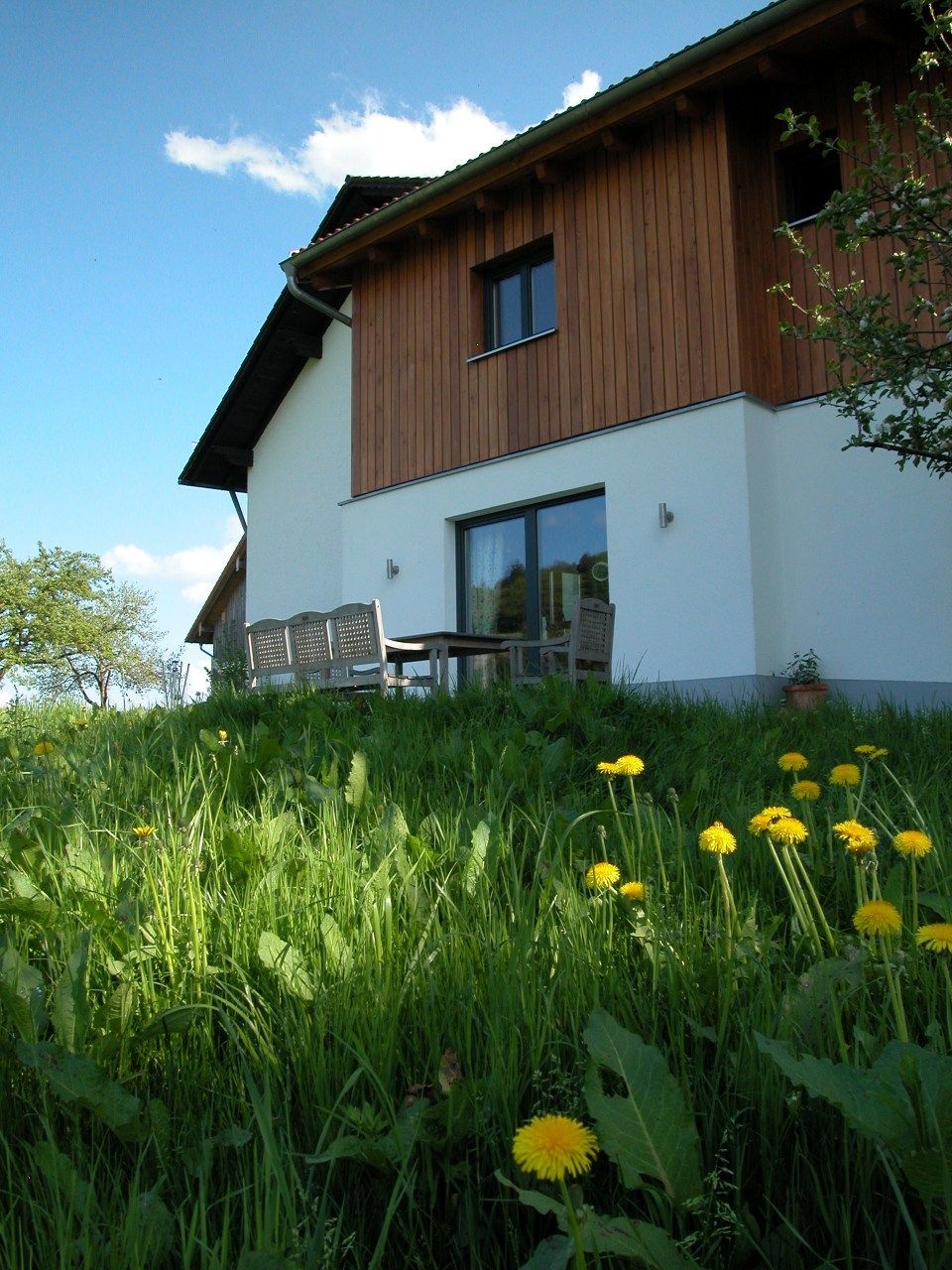 Ein weißes Haus mit einem Holzdach ist von hohem Gras und gelben Blumen umgeben. Foto von Eder Christiane Kosmetikstudio