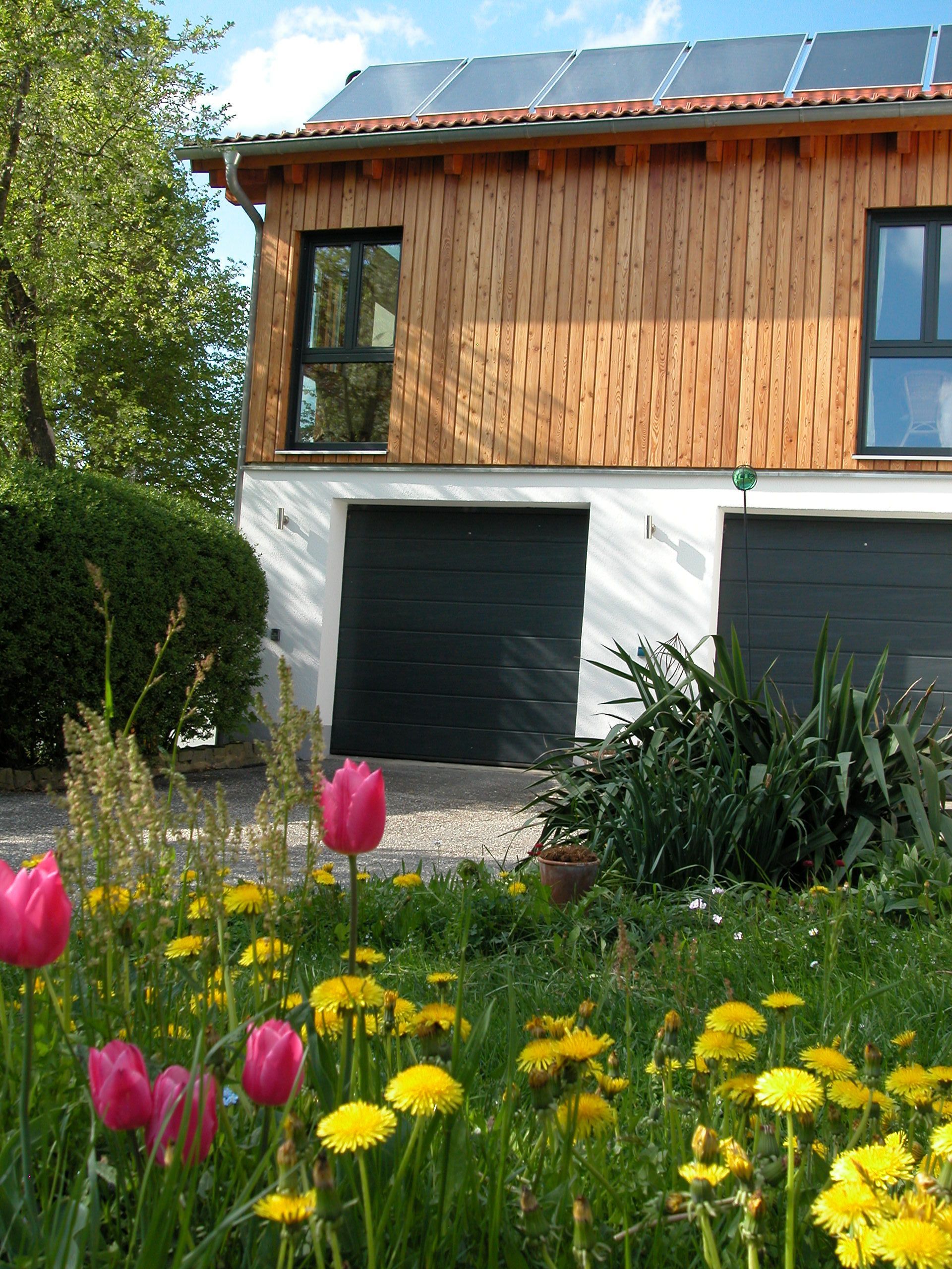 Ein weißes Haus mit einem Holzdach ist von hohem Gras und Blumen umgeben. Foto von Eder Christiane Kosmetikstudio