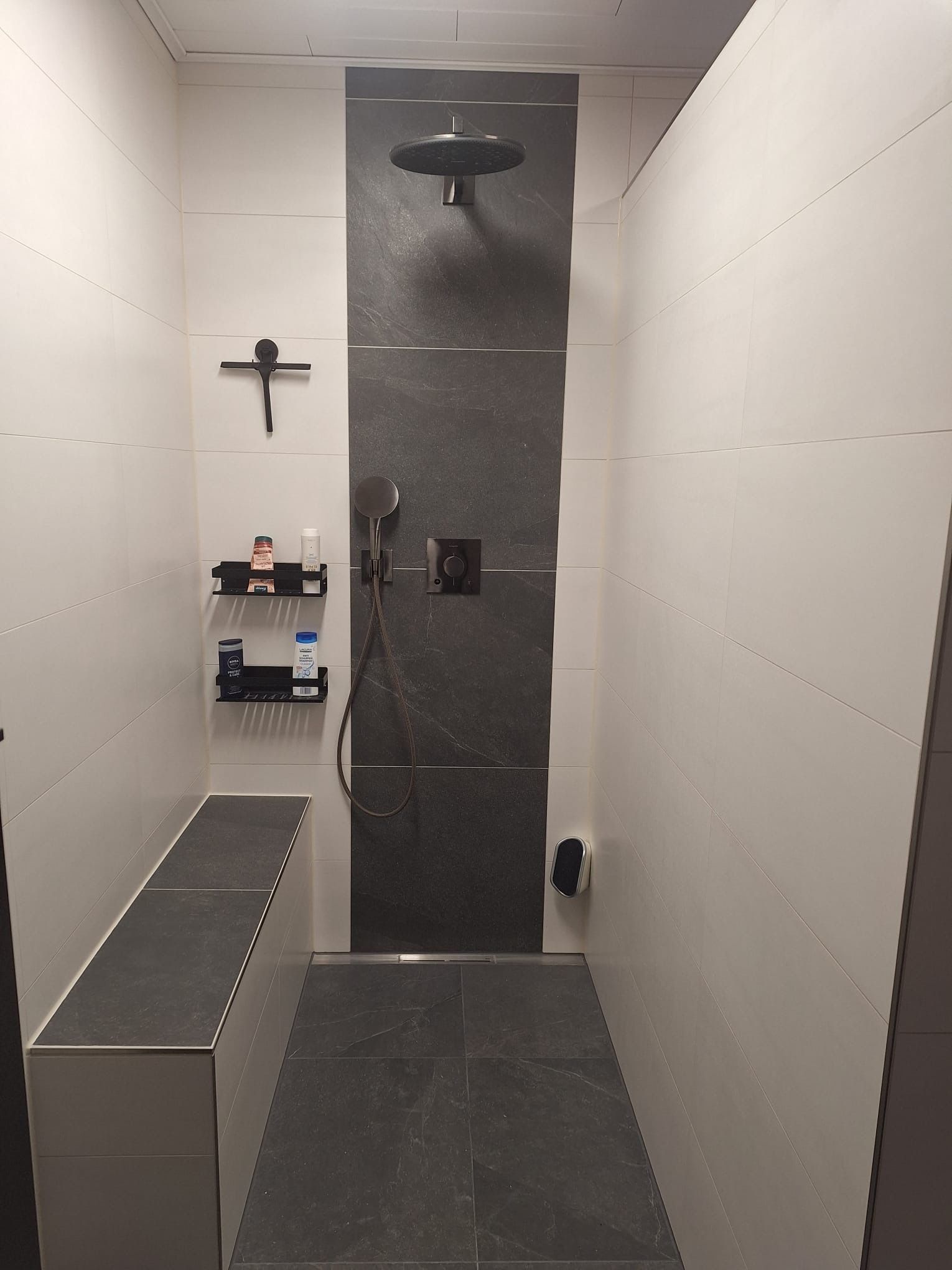 Schmale, geflieste Dusche mit schwarzen Armaturen, Wandregal und Sitzbank in einem modernen Badezimmer