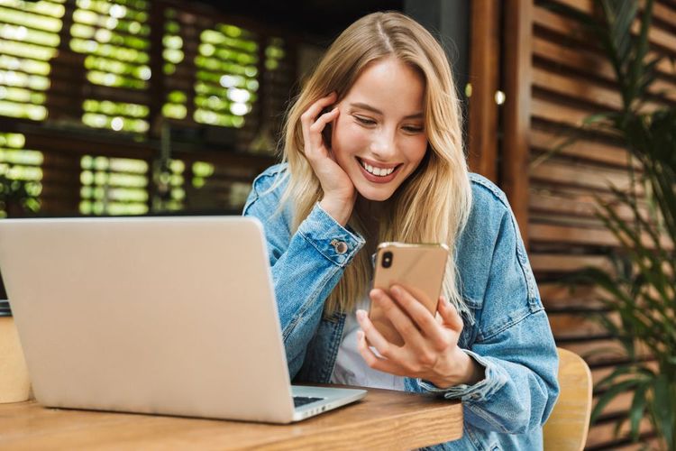 Una persona con una chaqueta vaquera sonríe mientras sostiene un teléfono inteligente, sentada en una mesa de madera con un ordenador portátil abierto.