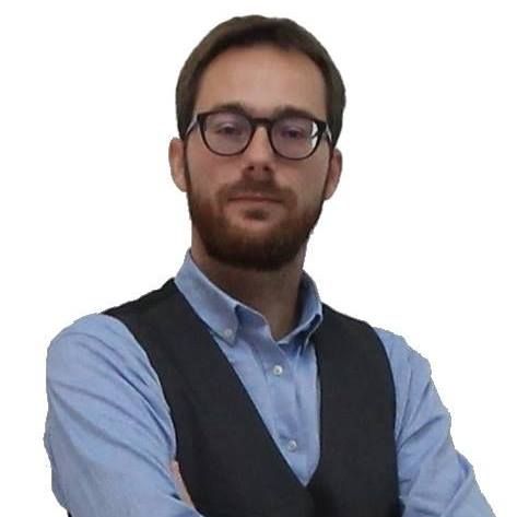 Un hombre con barba y gafas lleva una camisa azul y un chaleco negro.