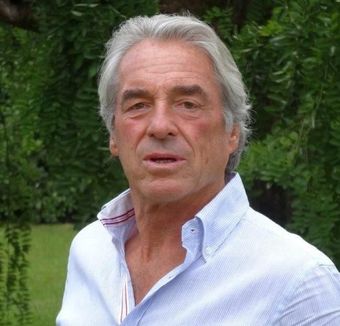 Portrait en buste d'un homme en chemise bleu clair, debout à l'extérieur devant un feuillage vert.