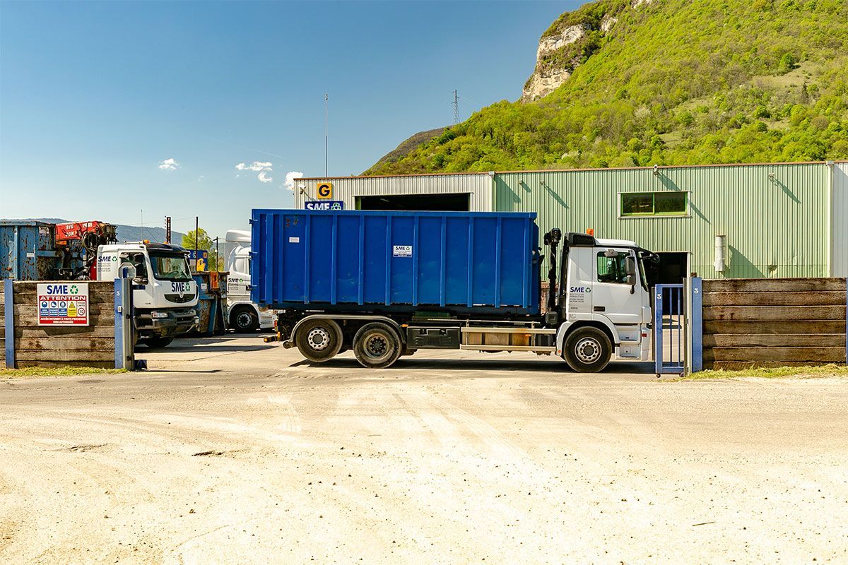 Un camion-benne bleu dans une recyclerie à côté d'une colline verdoyante, avec des pompes et un petit camion blanc à proximité.