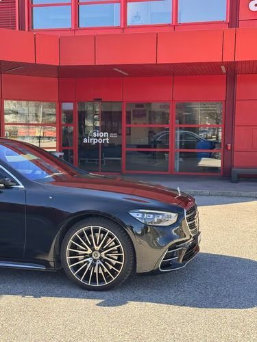 Une Mercedes-Benz sombre garée devant un bâtiment rouge avec un panneau « aéroport » sur l'entrée vitrée.