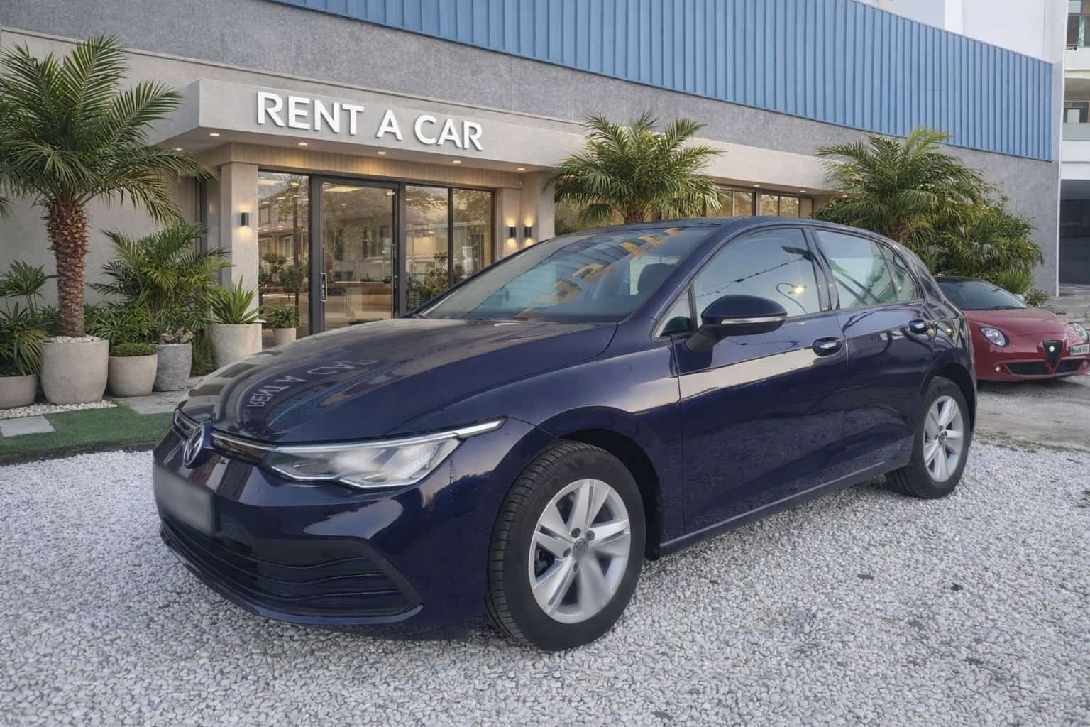 Un coche compacto azul oscuro aparcado frente a un moderno negocio de alquiler de coches sobre una superficie de grava.