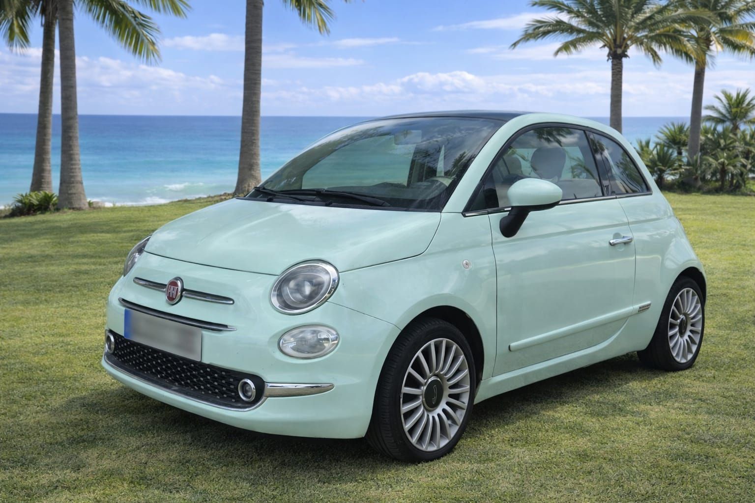 Un Fiat 500 de color verde claro estacionado en un césped con palmeras y el océano al fondo.