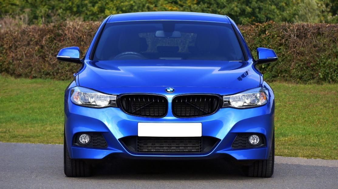 Vista frontal de un sedán BMW azul estacionado en una carretera con césped y setos detrás.