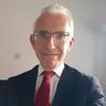 Homme en costume sombre, cravate rouge et lunettes, souriant devant un fond clair uni