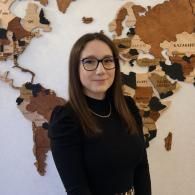 Une femme portant des lunettes se tient devant une fresque représentant une carte du monde, vêtue d'un haut noir et souriant doucement.