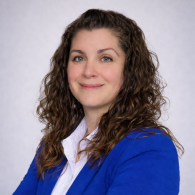 Femme en blazer bleu et chemise blanche, souriant sur fond gris clair