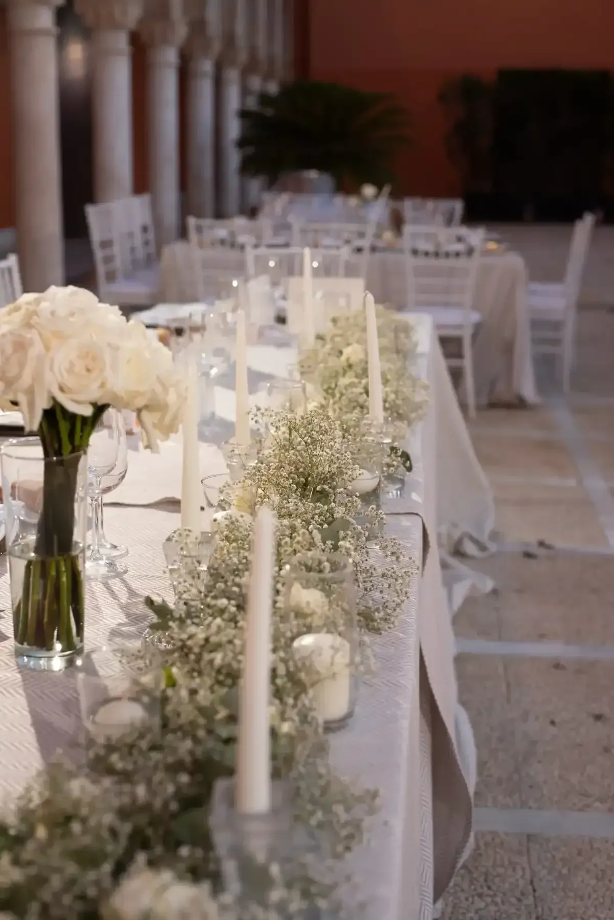 Mesa de recepción nupcial blanca con flores, velas y vegetación en un lugar con iluminación tenue.
