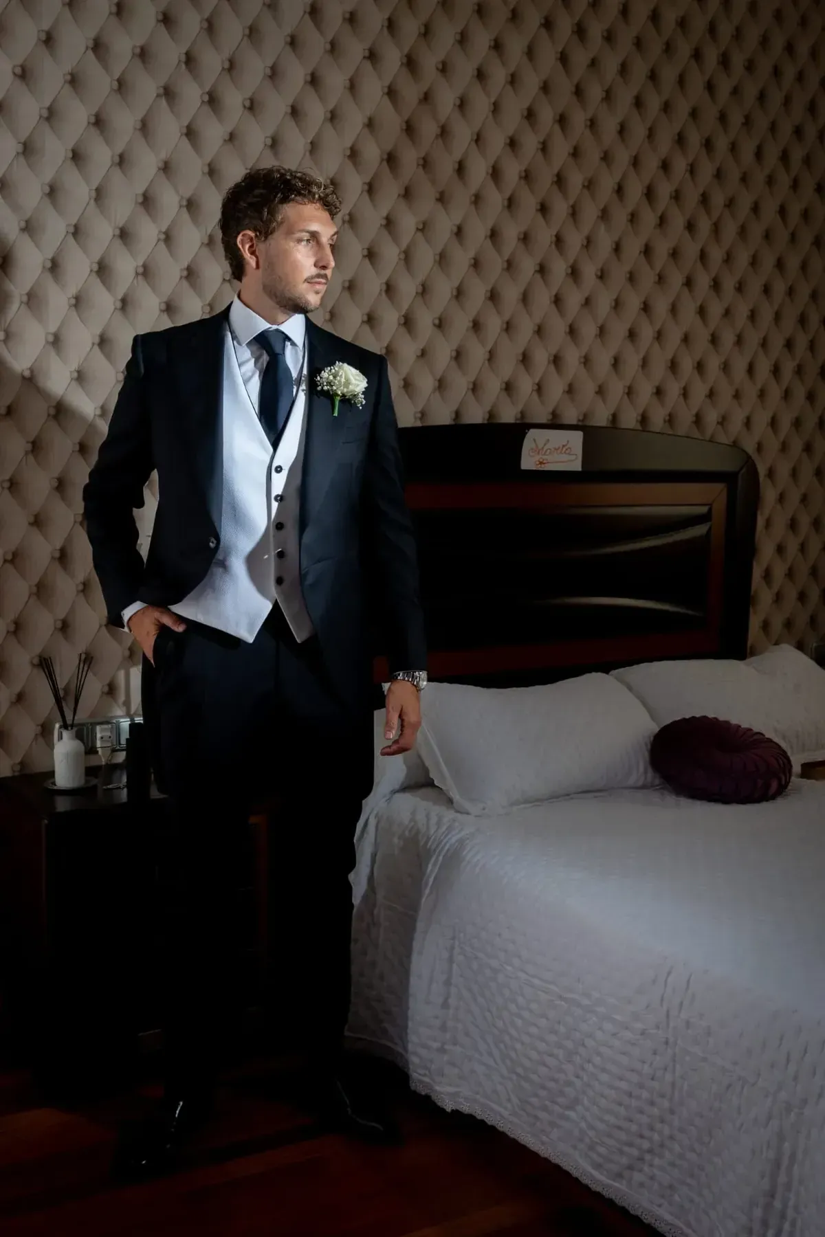 Un hombre con un traje oscuro está de pie junto a una cama en una habitación de hotel con iluminación tenue.