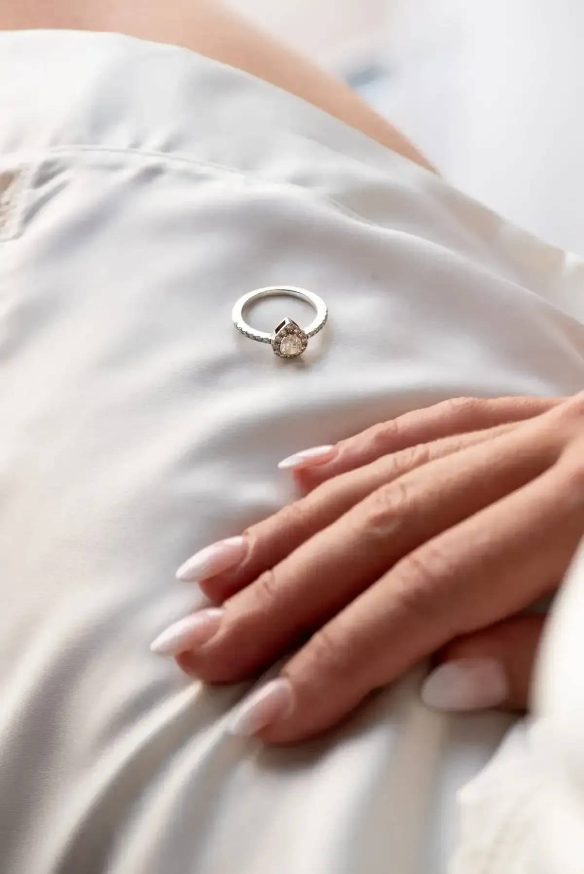 Anillo de plata sobre satén blanco junto a una mano en reposo con uñas largas color nude.