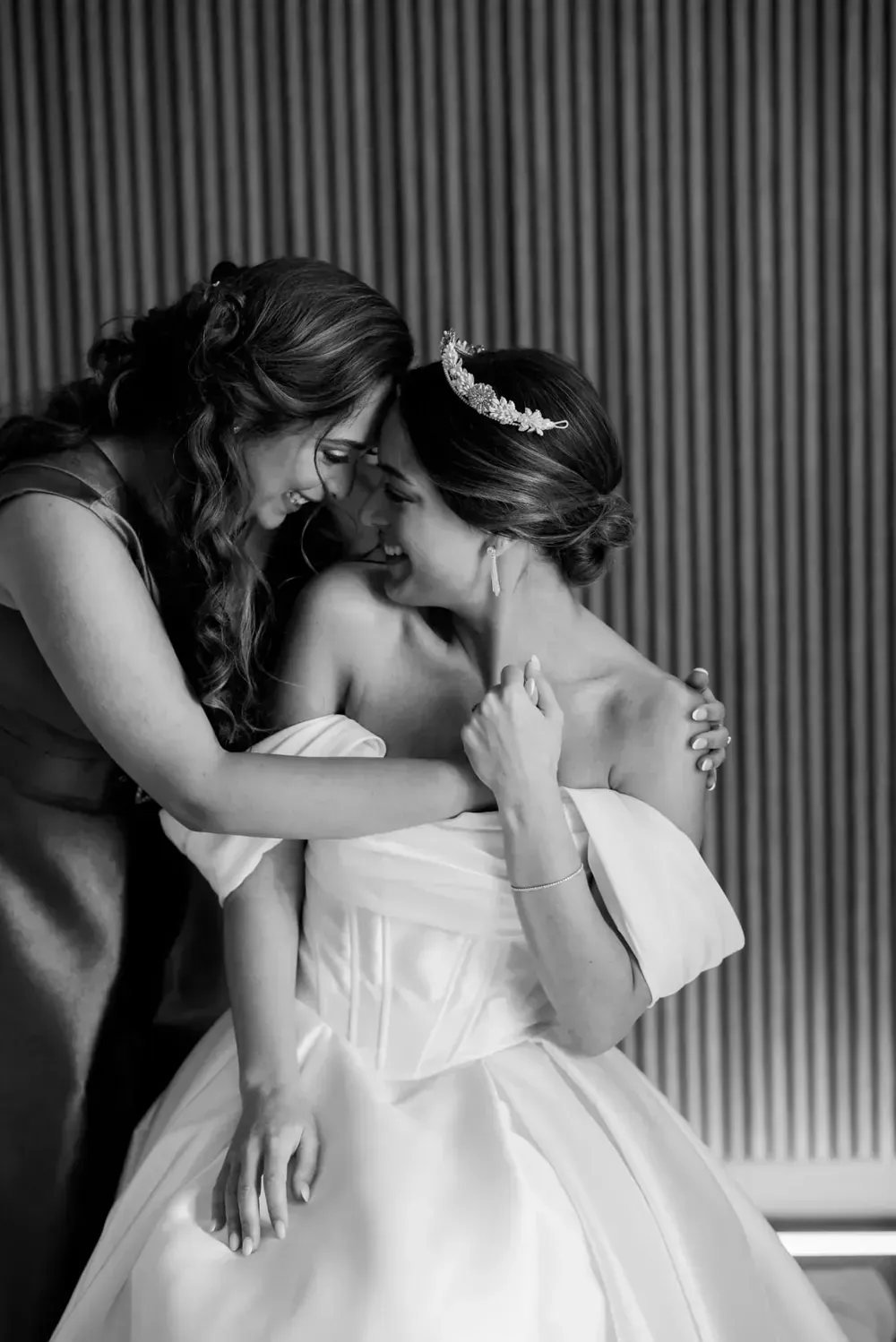 Dos mujeres se abrazan vestidas de gala; una de ellas lleva un vestido con los hombros descubiertos y un tocado floral.