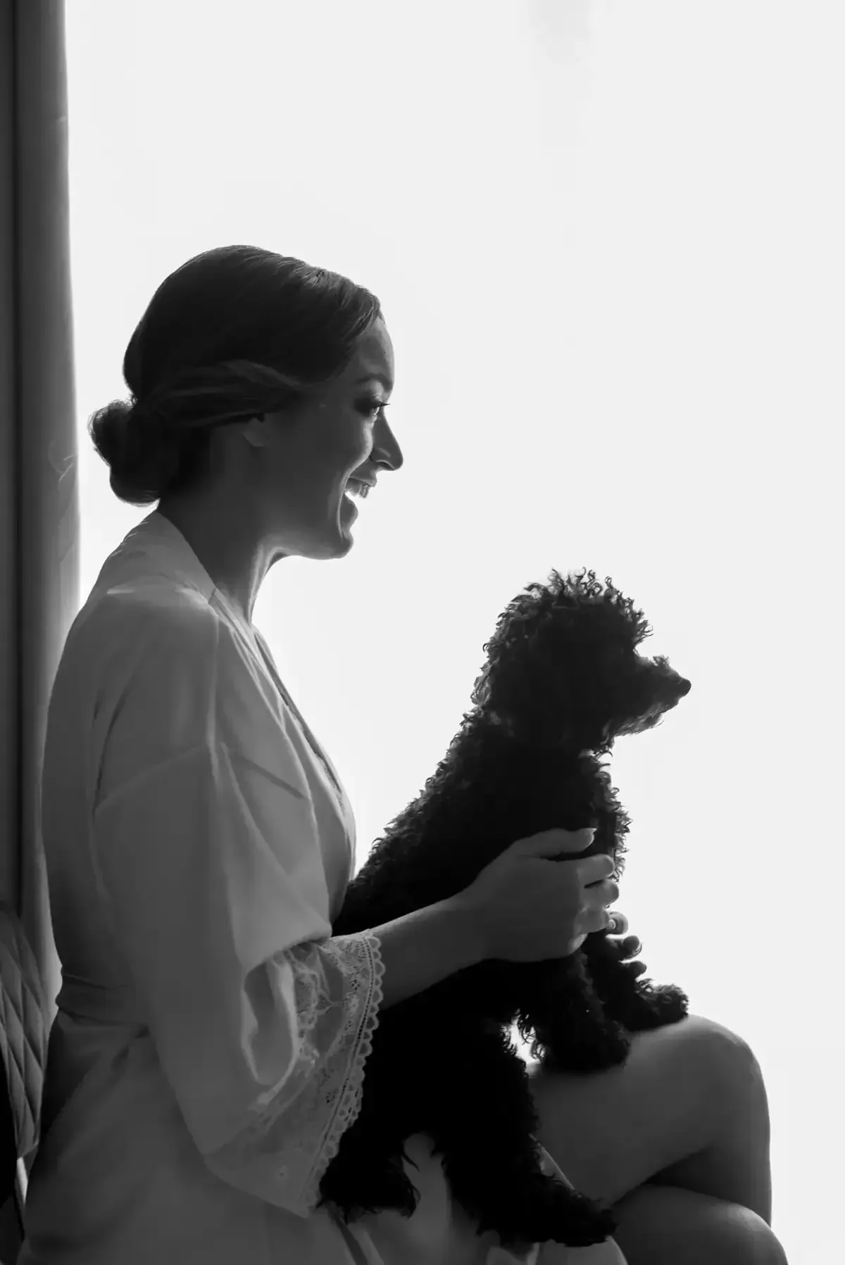 Silueta de una mujer sentada junto a una ventana luminosa, con un perrito en su regazo.