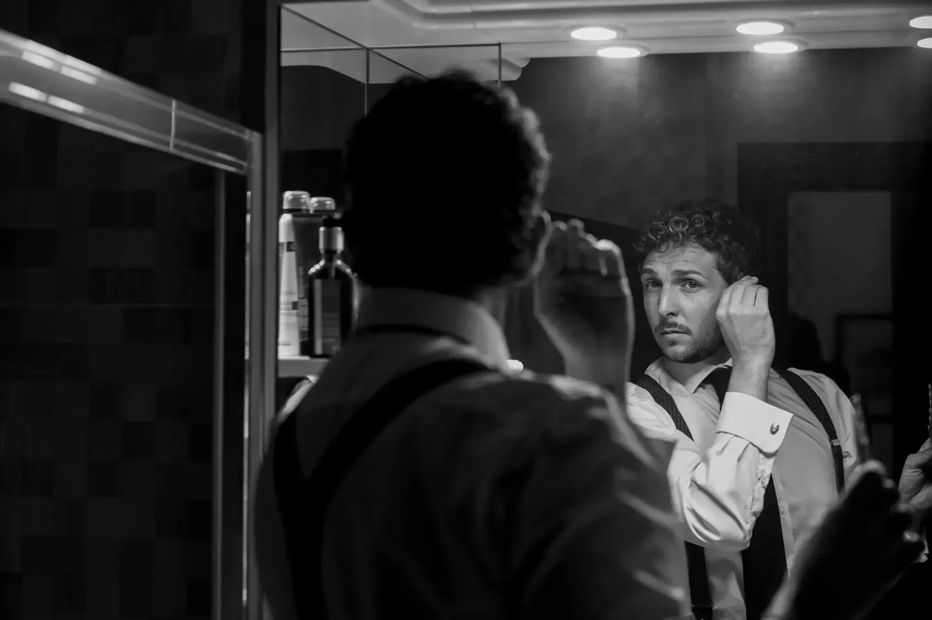 Un hombre se ajusta la corbata y el cuello de la camisa frente al espejo del baño; la imagen se ve en blanco y negro.
