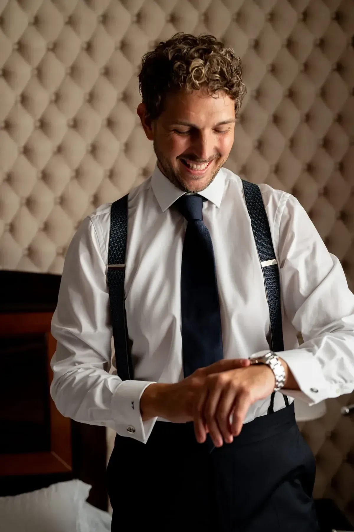 Hombre sonriente con camisa blanca y tirantes mirando su reloj en una habitación acolchada.