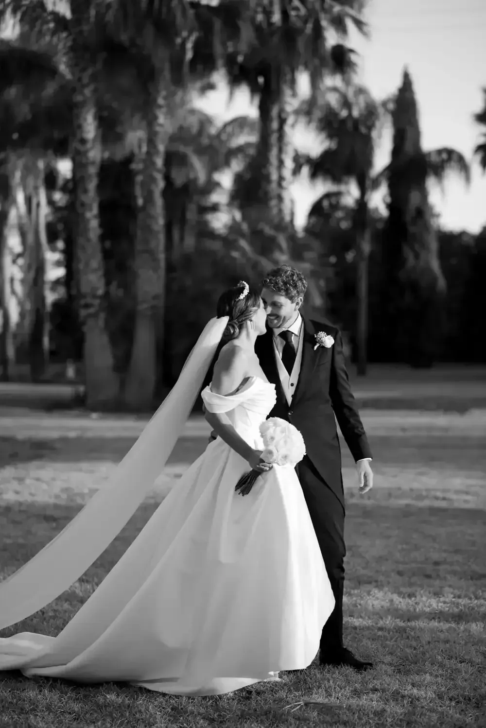 Novios de pie en un césped con palmeras al fondo, tomados de la mano y besándose.