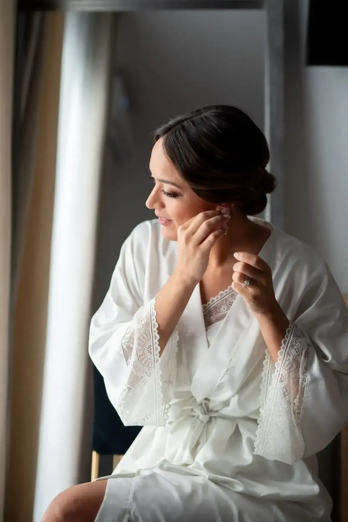 Mujer con bata blanca ajustándose un pendiente junto a una ventana con luz natural tenue.