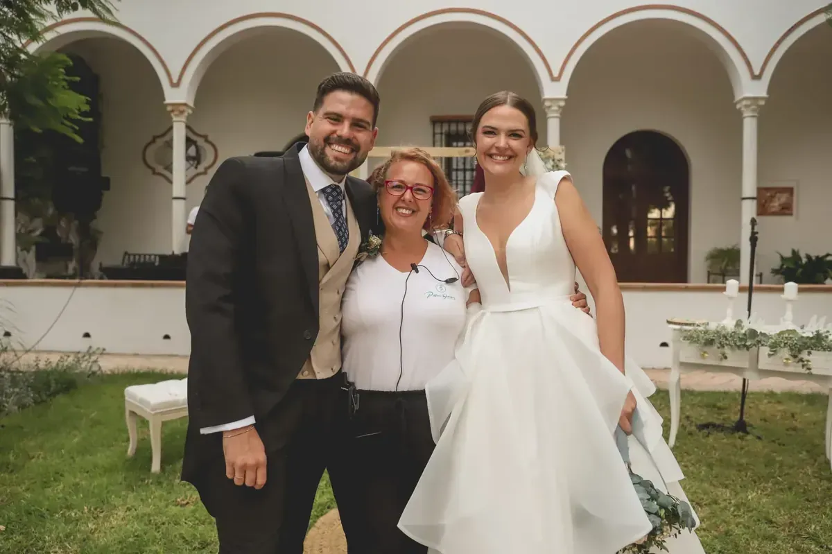 Tres personas sonrientes posan al aire libre frente a un patio arqueado; una de ellas lleva un vestido de novia blanco.