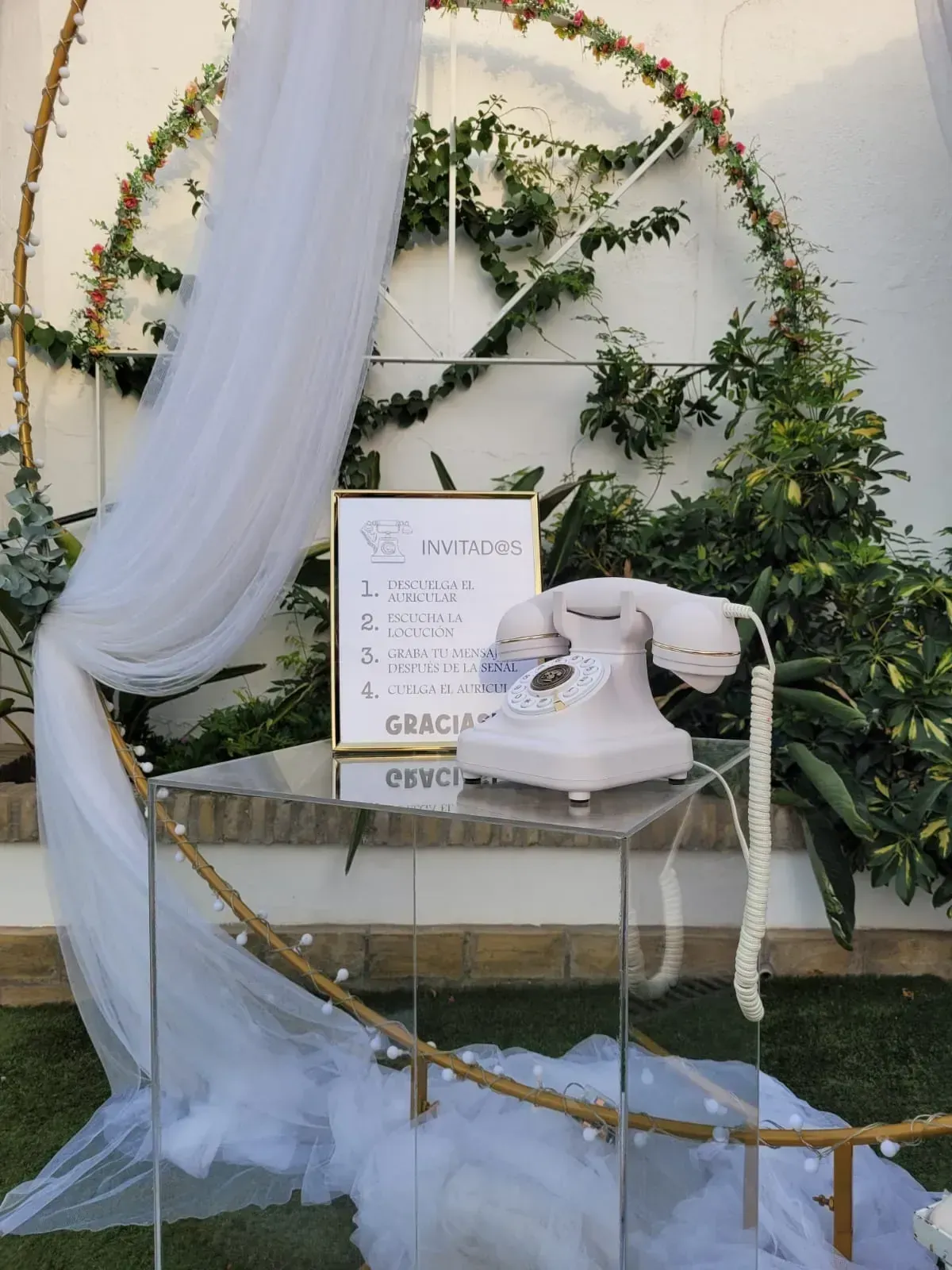 Un teléfono blanco de disco sobre una mesita con un cartel, sobre un fondo floral y una tela blanca drapeada.
