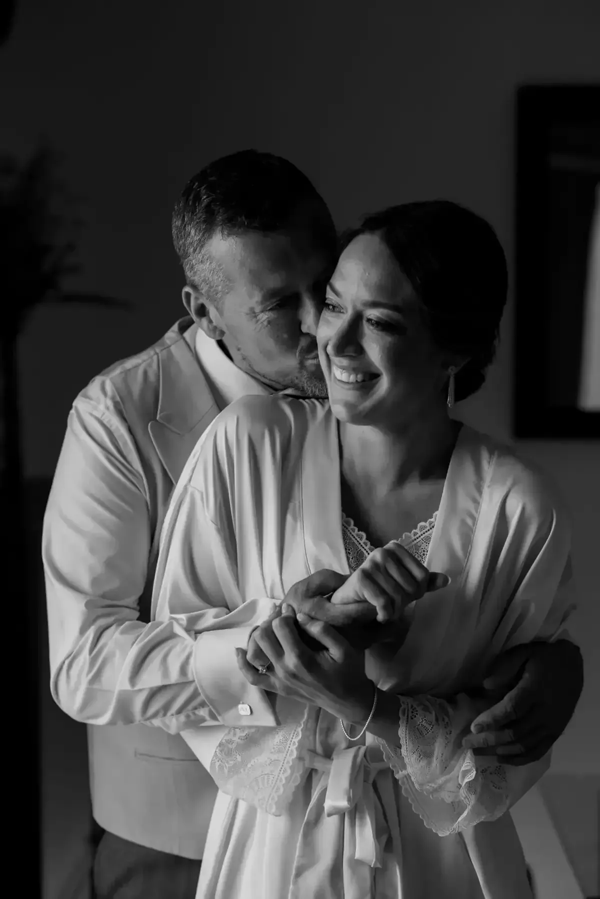 Una pareja se abraza en una habitación con poca luz, sonriendo y tomados de la mano cerca de una ventana.